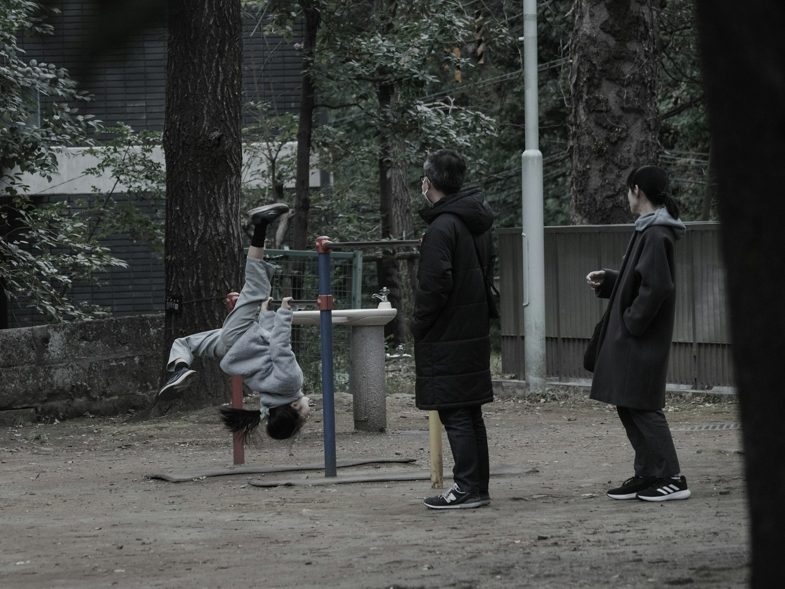 Enfant jouant sur des équipements de parc avec des adultes regardant dans un parc entouré d'arbres