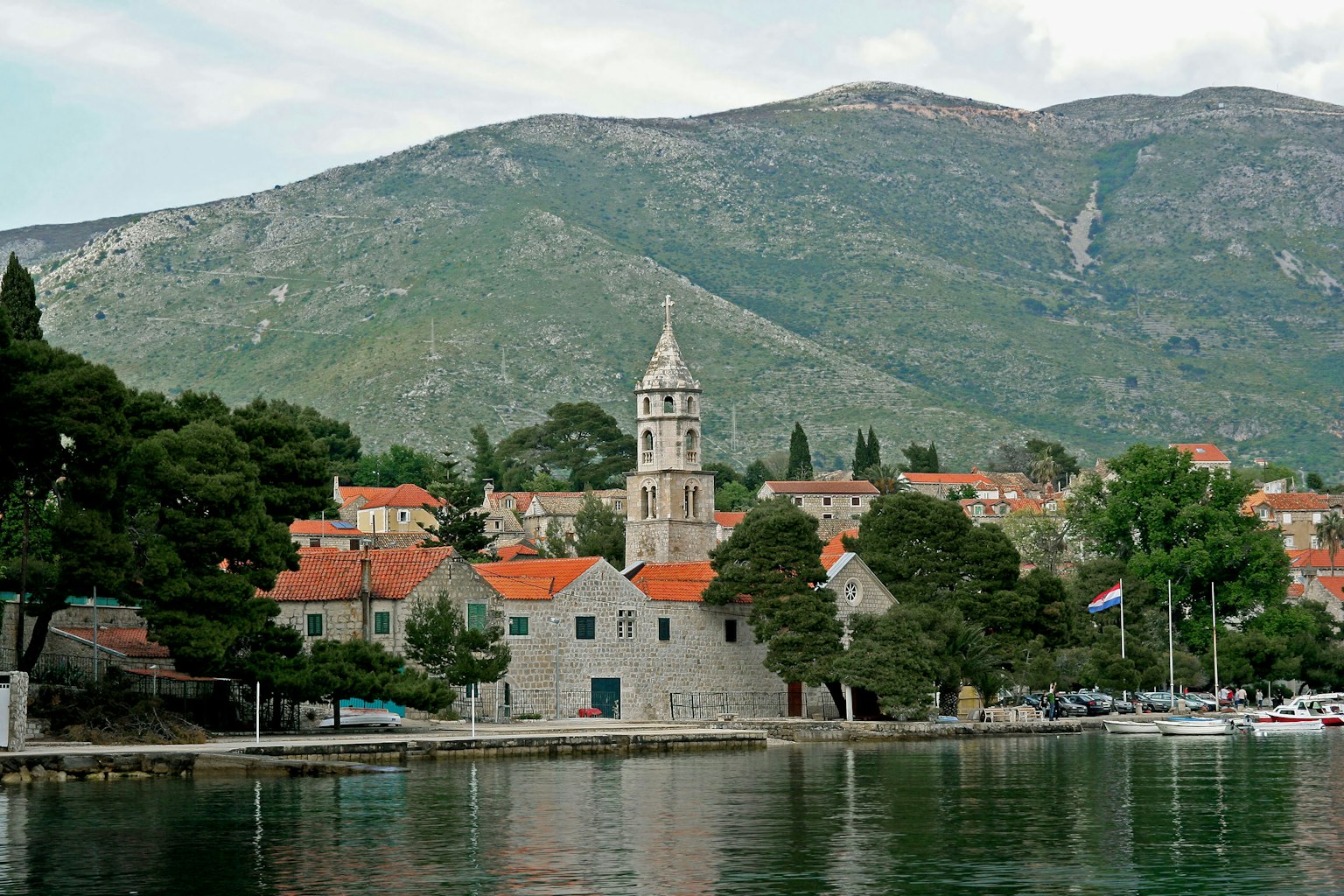 Scenic view of a quiet coastal town featuring stone buildings with orange rooftops surrounded by green trees and mountains