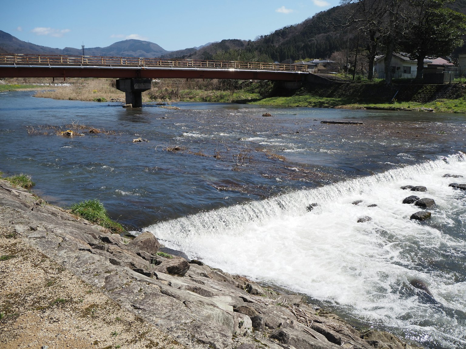 Malersicher Blick auf einen Fluss mit einer Brücke, sanfter Fluss und Wasserfall, blauer Himmel und Berge