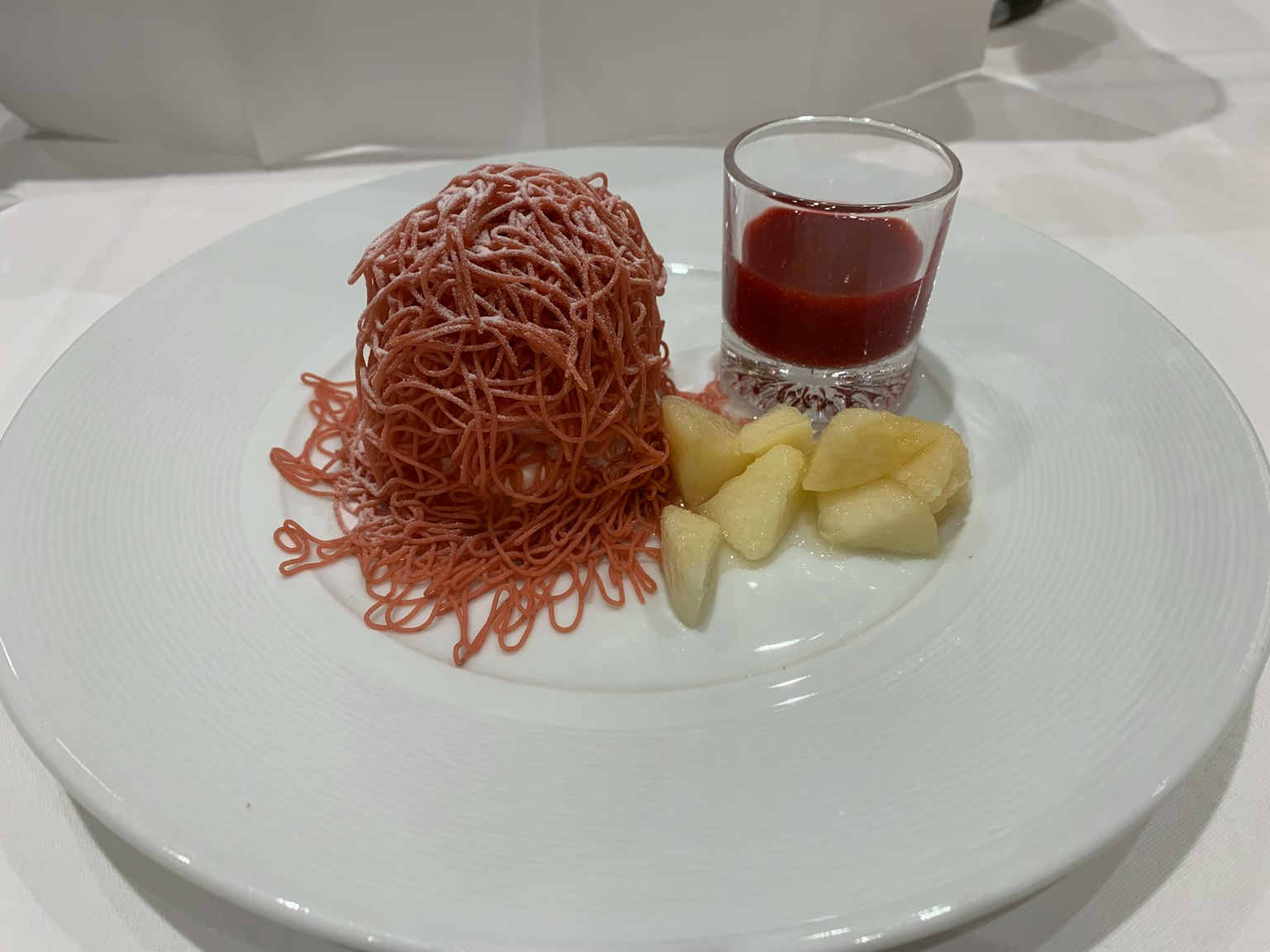 Dessert su un piatto bianco con crema rosa a forma di noodle un piccolo bicchiere di salsa rossa e fette di mela