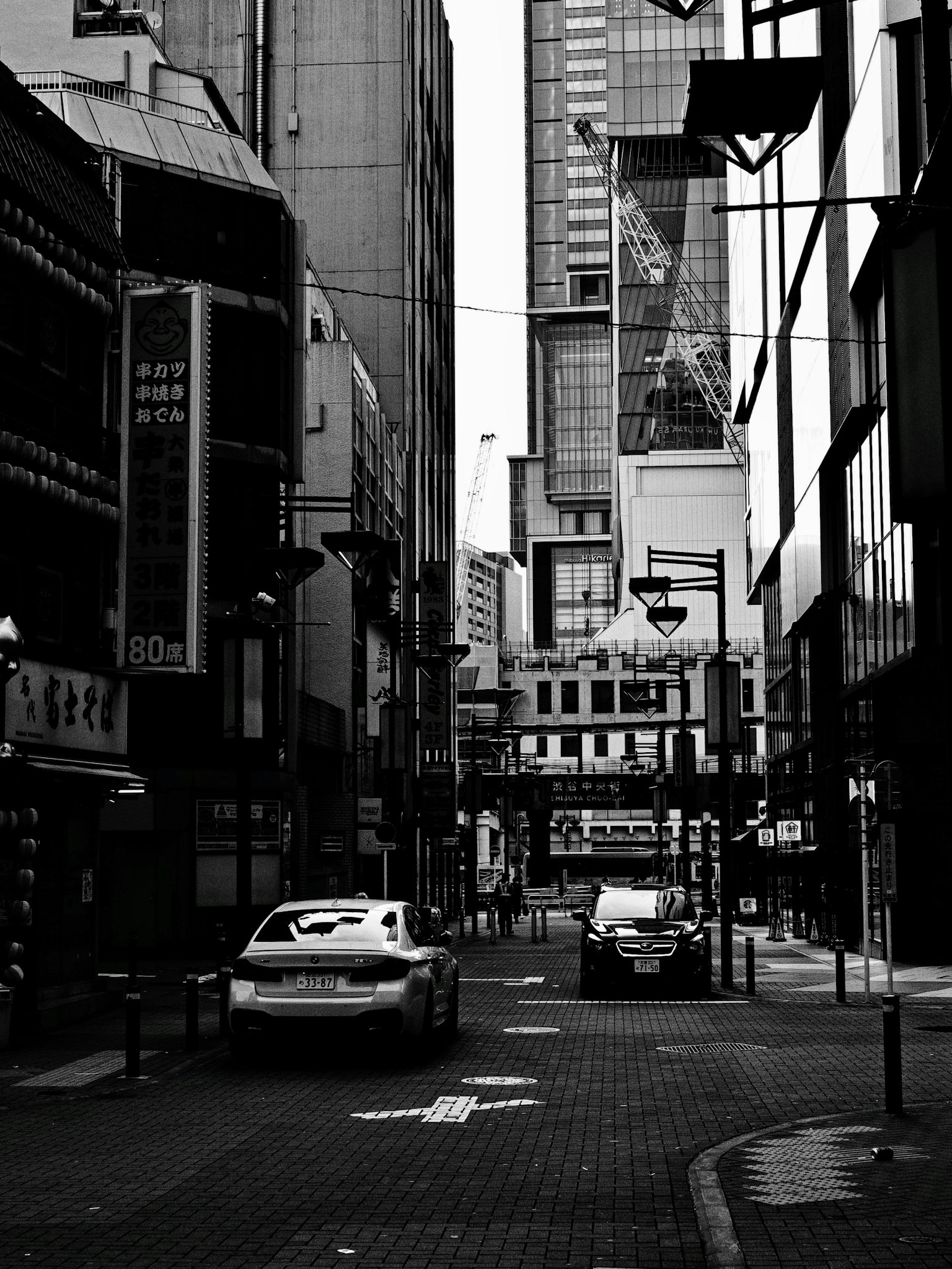 ทิวทัศน์เมืองขาวดำที่มีถนนแคบและรถยนต์อยู่ระหว่างตึกสูง