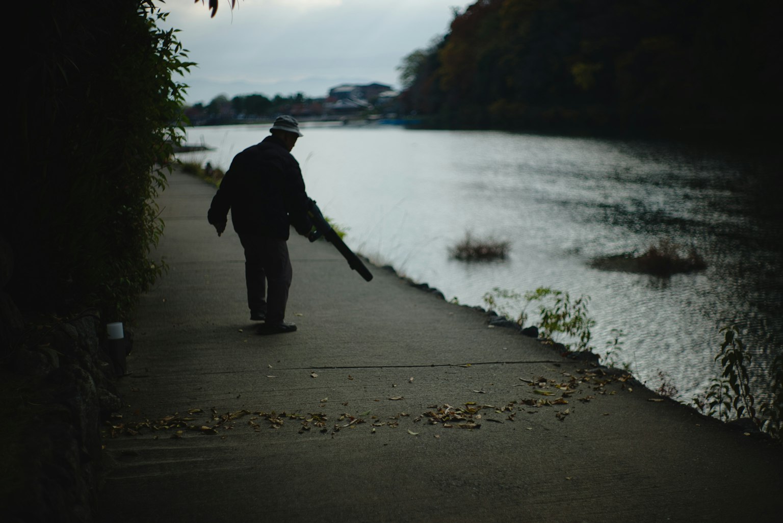 川沿いを歩く男性のシルエット 落ち葉が散らばる歩道 曇り空の下の静かな風景