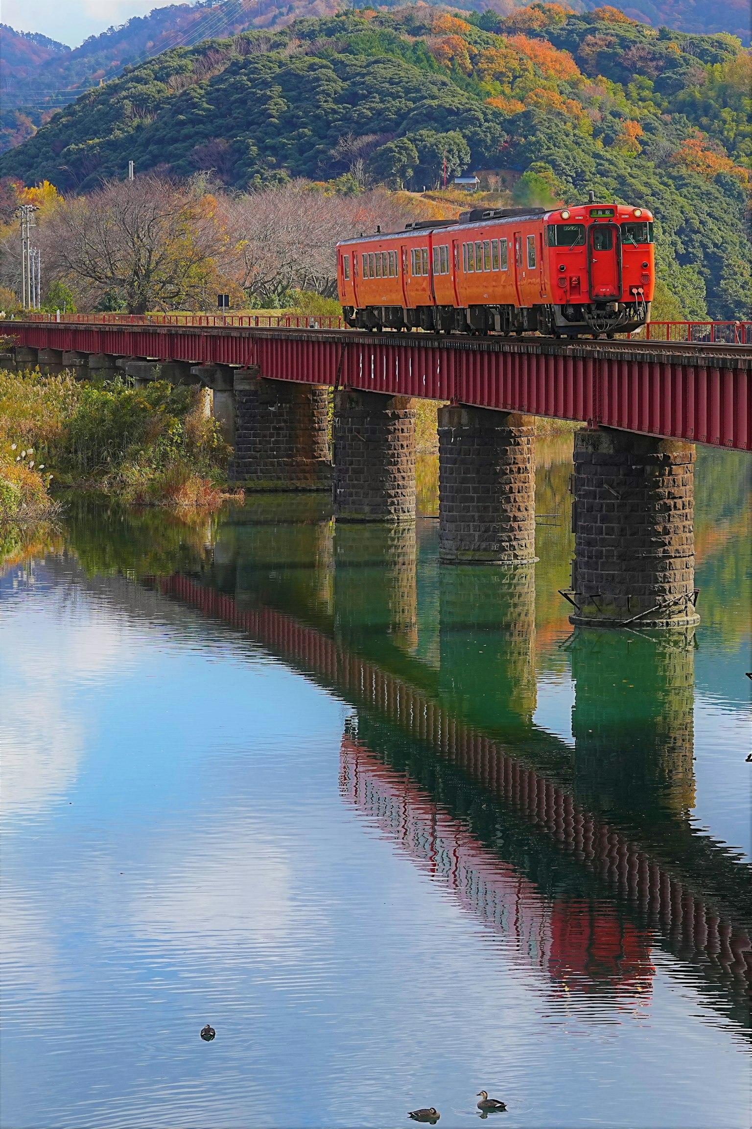 Un tren rojo cruzando un puente con un reflejo en el río