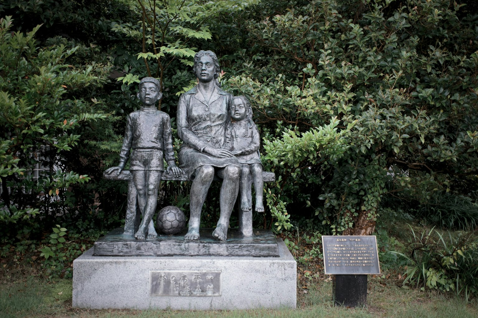Bronzestatue einer Frau, die mit Kindern sitzt, umgeben von Grün