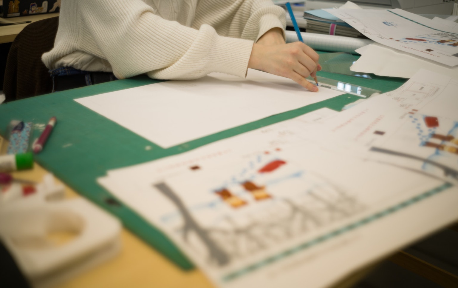 A person drawing with a pencil on a blank sheet surrounded by colorful art supplies and sketches