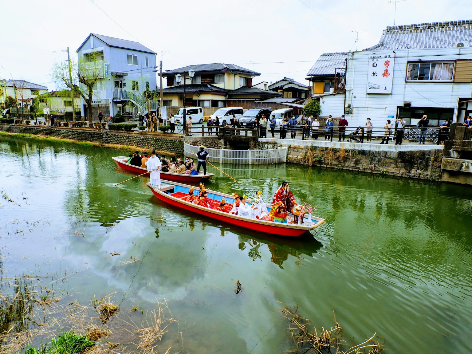 Perahu merah berlayar di sungai hijau dengan rumah tradisional di latar belakang