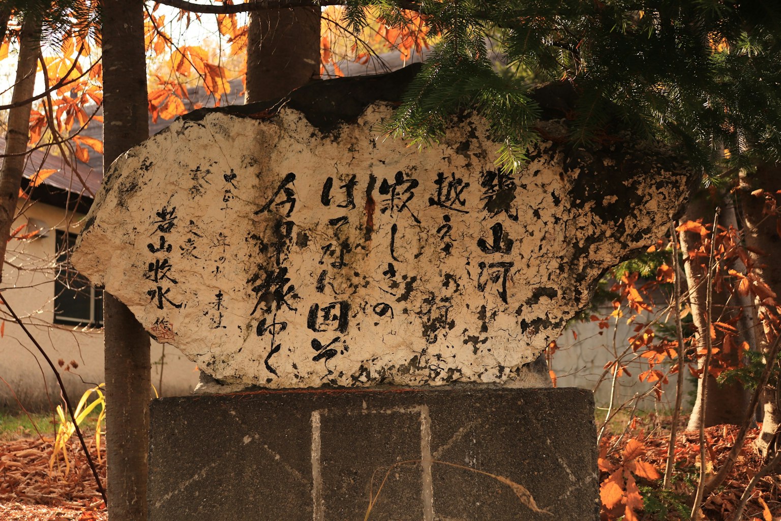 一块古老的石碑，周围环绕着秋天的风景，刻有日文
