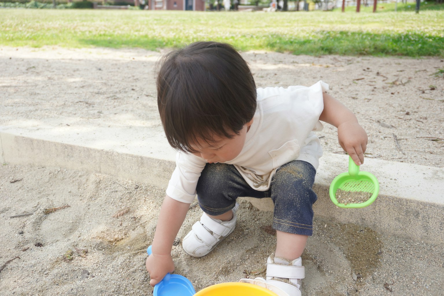 Anak bermain di kotak pasir mengenakan kaos putih dan celana biru menggali pasir