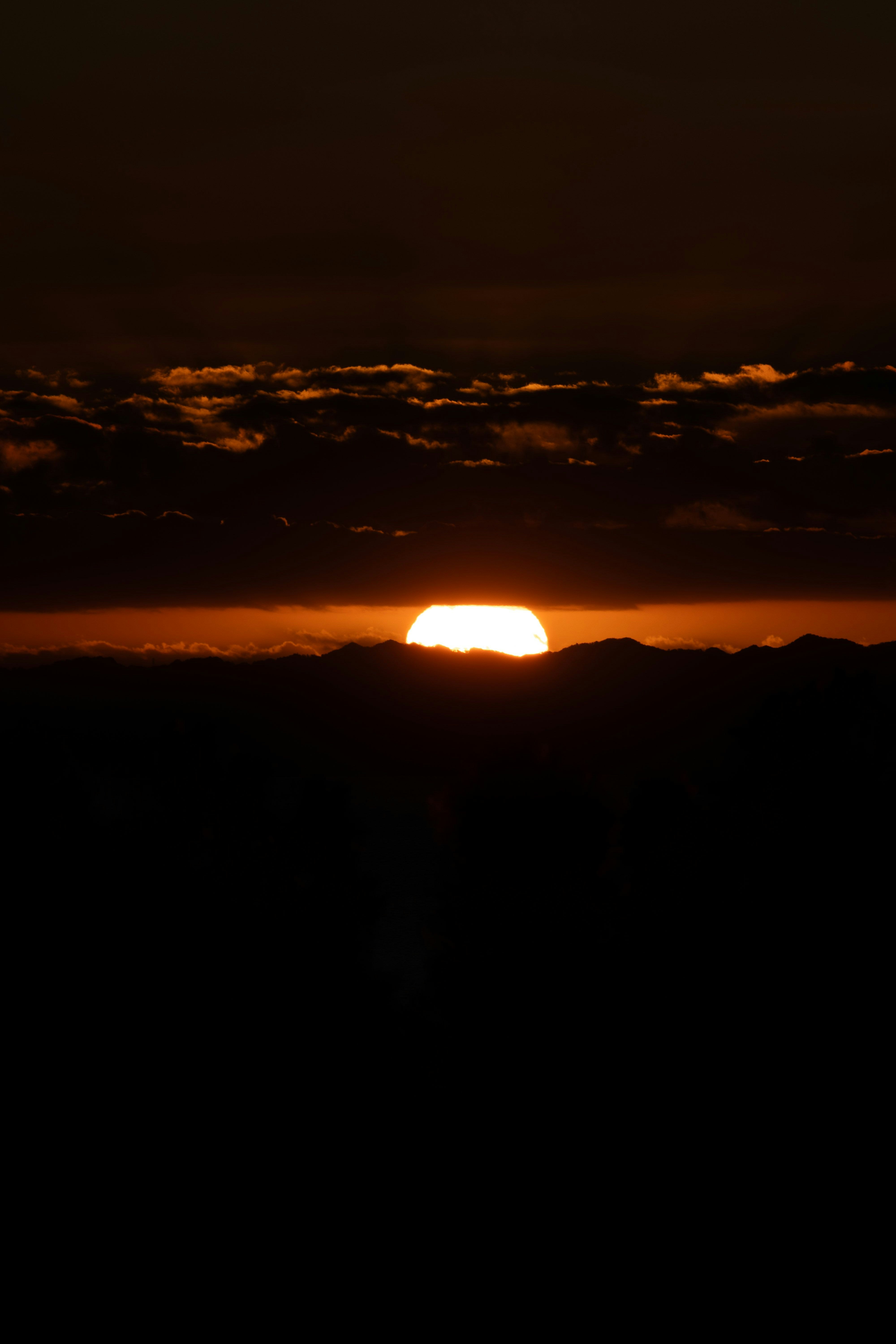山並みのシルエットと夕焼けの太陽が雲の間から差す風景
