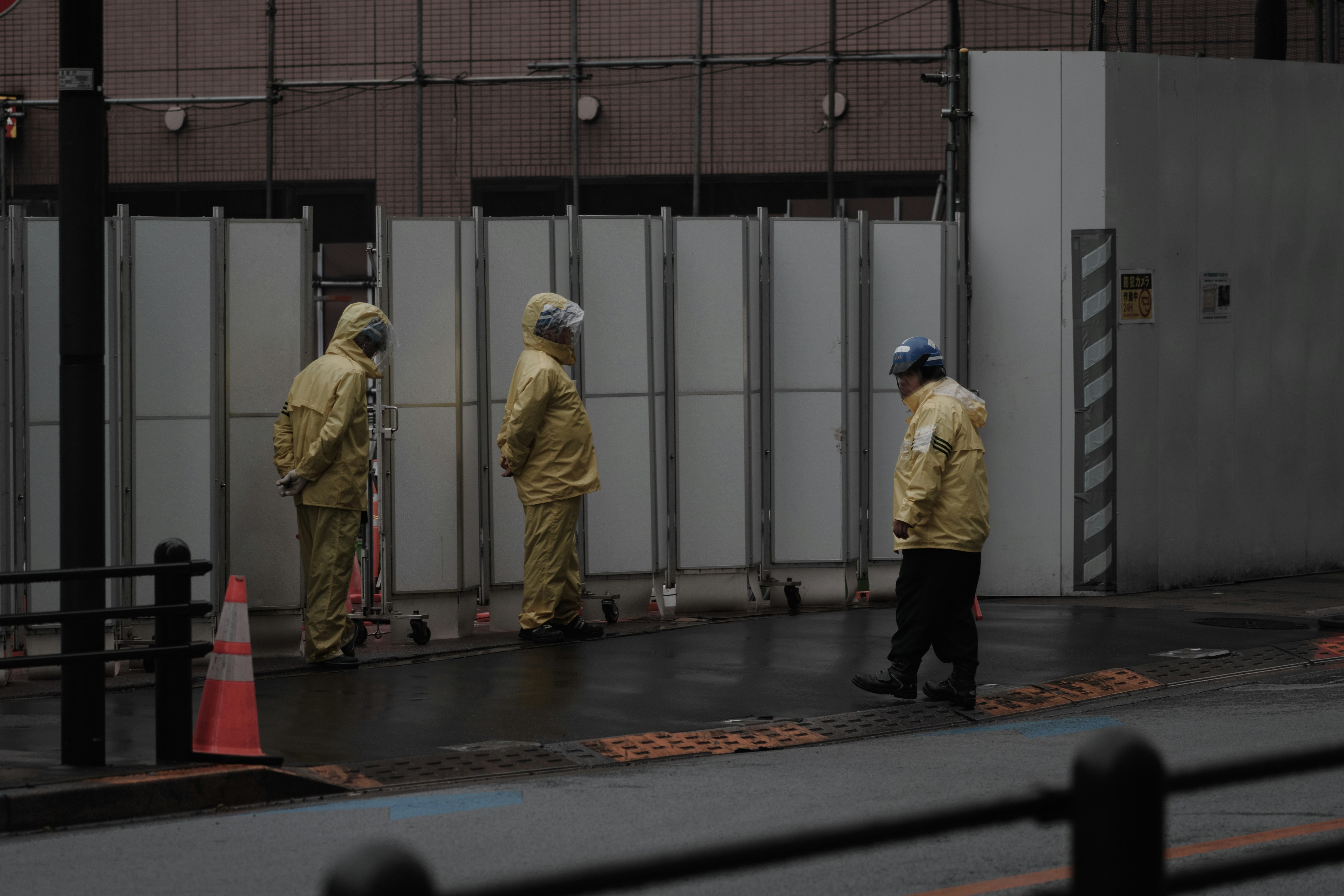 雨の路地で黄色い作業服の人が壁沿いに立つ東京の街並み