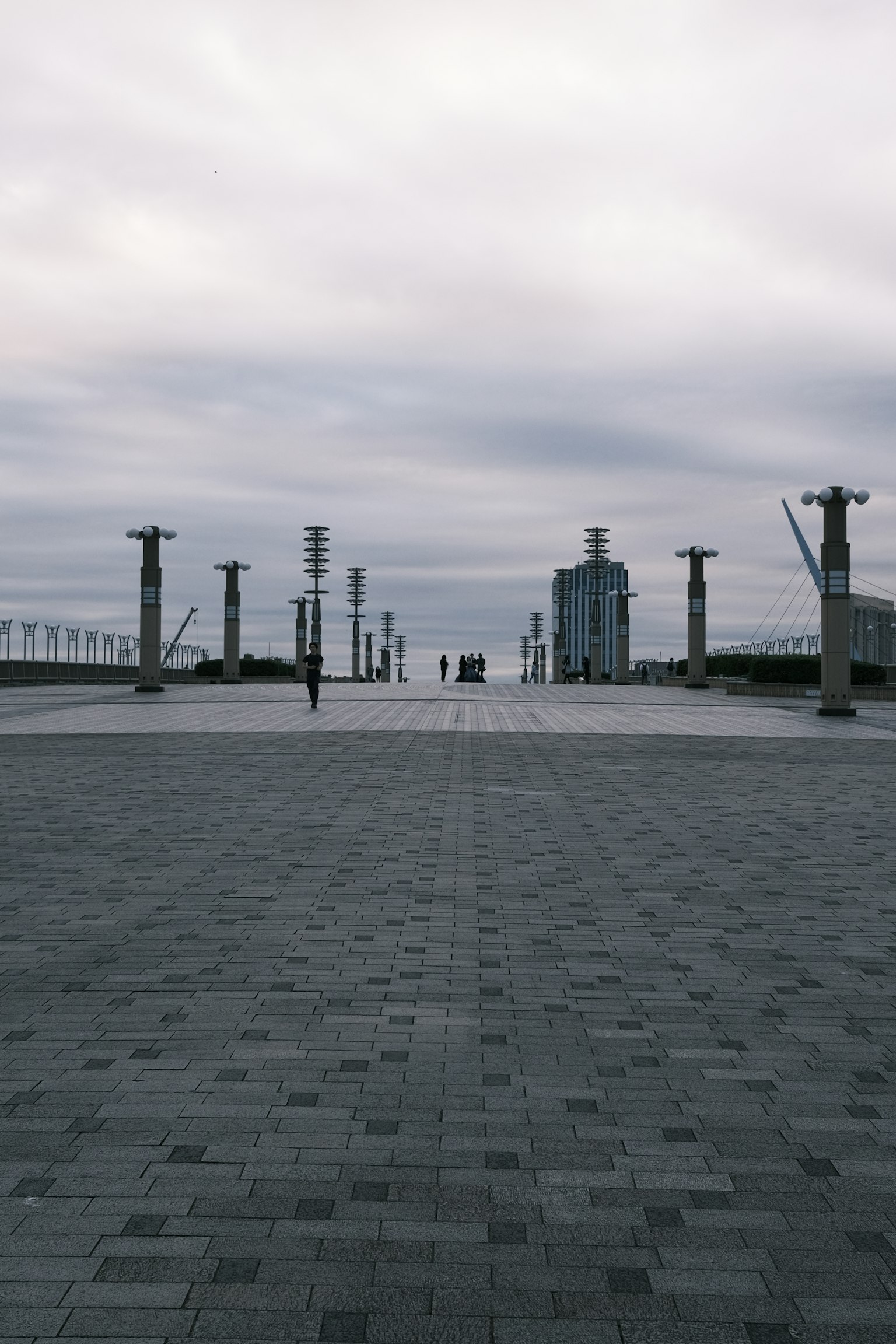 Wide paved area with people standing and buildings visible in the distance under a cloudy sky