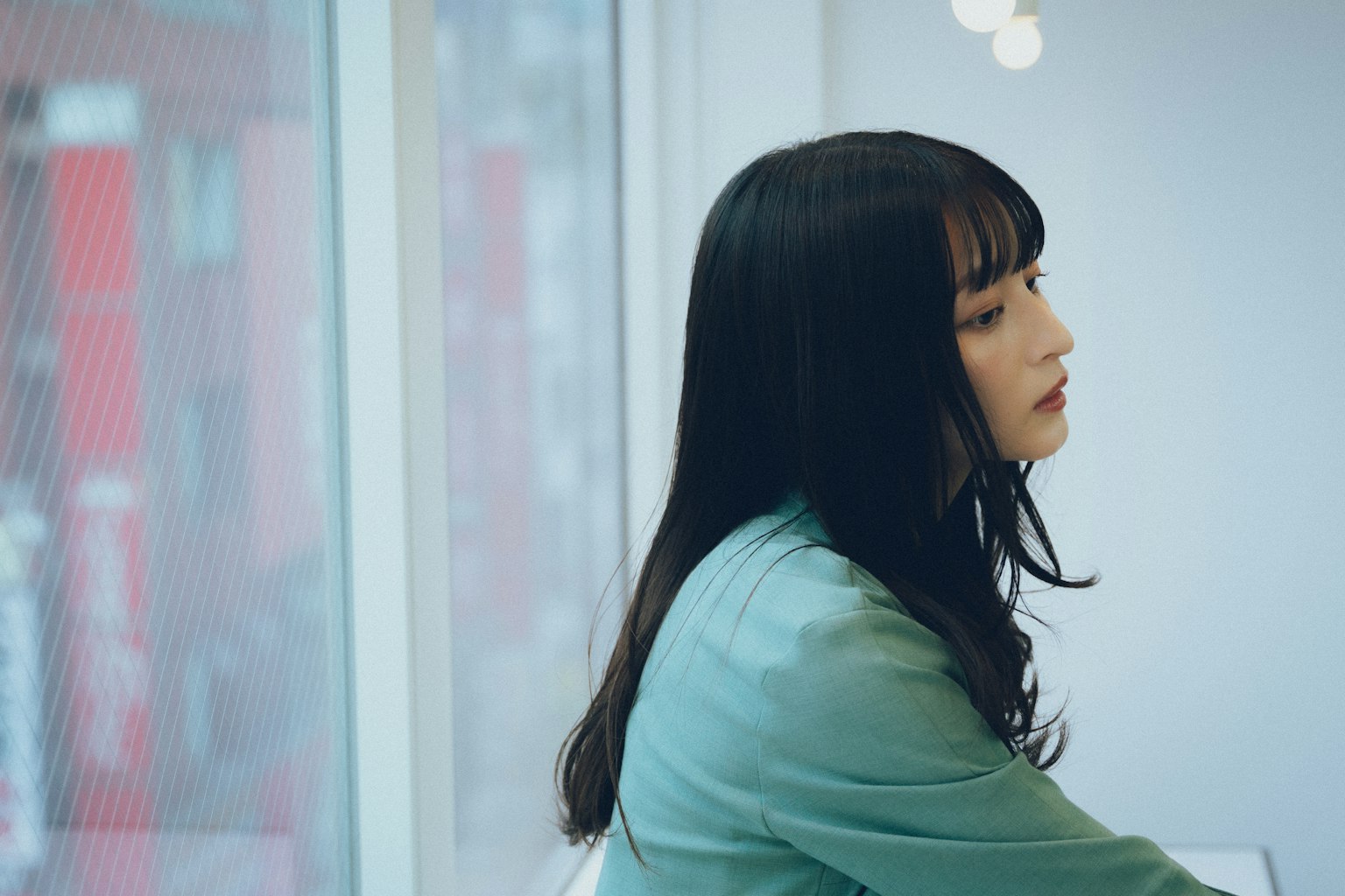 窗边深思的女性侧面图，穿着浅绿色衣服，长黑发，背景为模糊的红色建筑