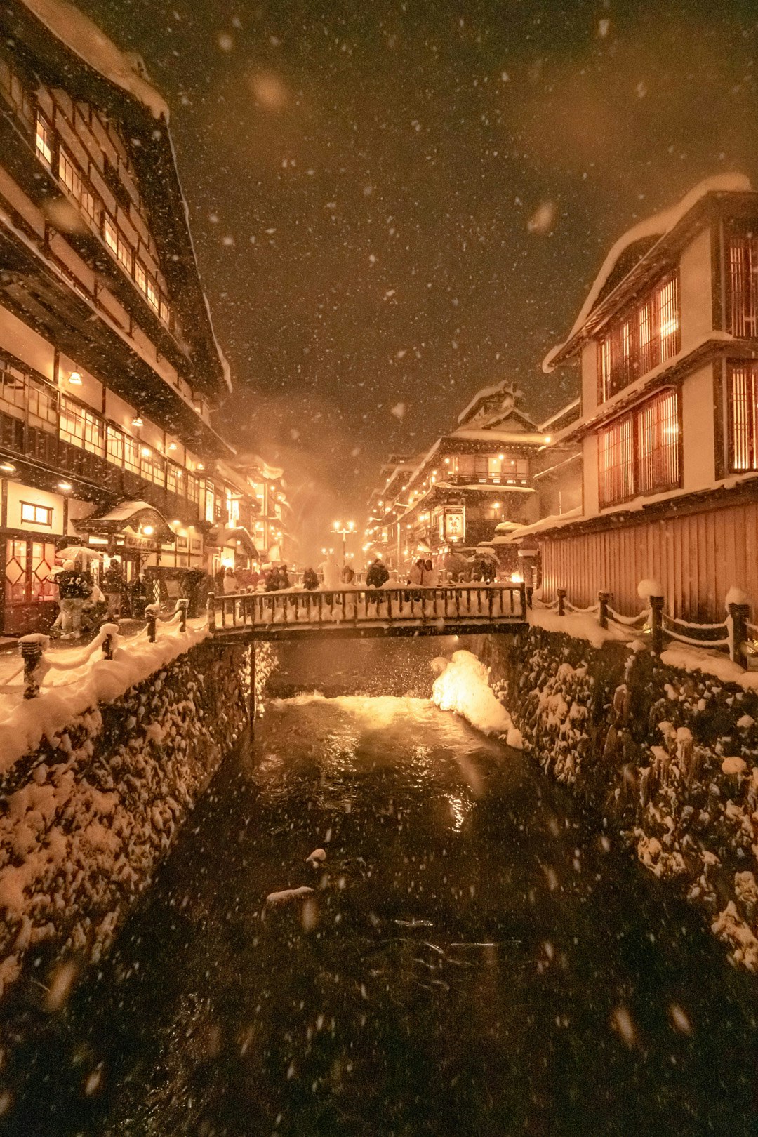 nighttime riverside street scene with falling snow and lantern lights