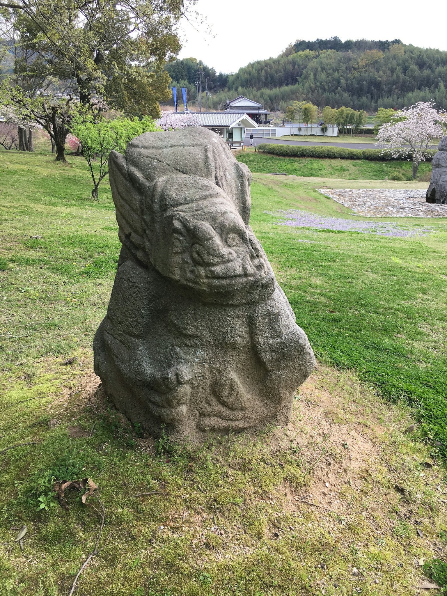 Steinskulptur auf grünem Gras mit Kirschbäumen im Hintergrund und einem kleinen Gebäude