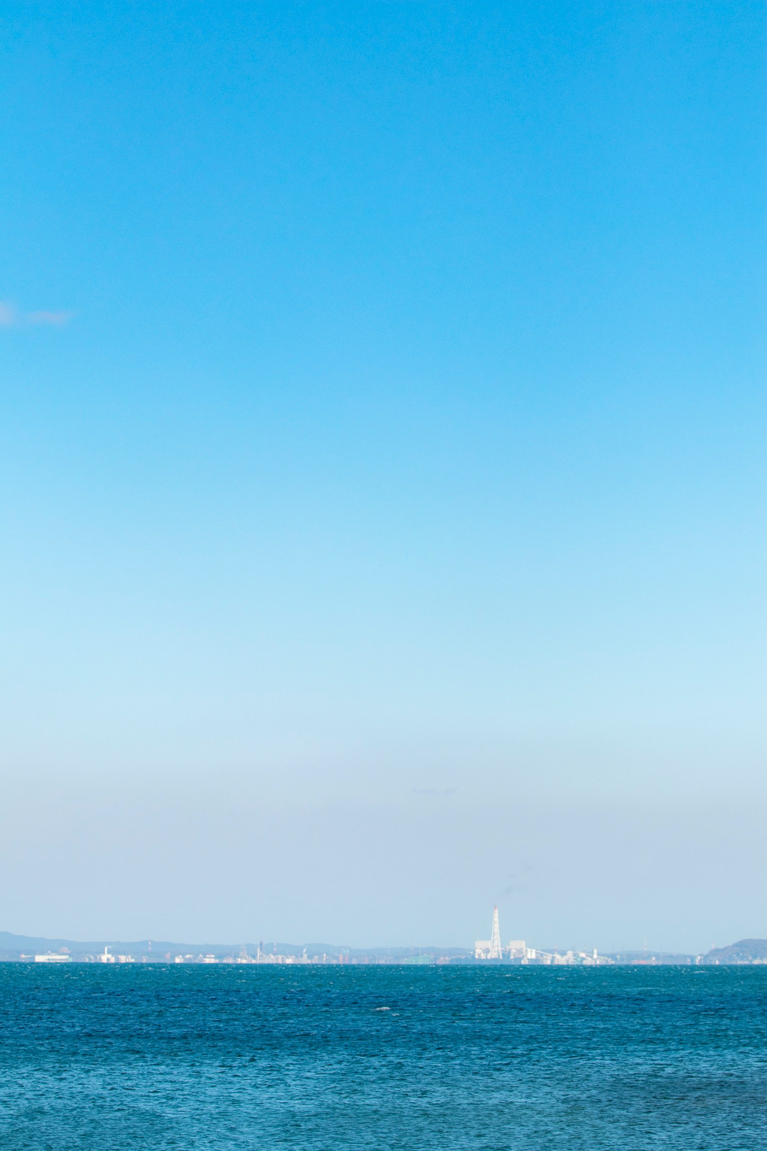 A landscape of blue sky and sea with a distant factory chimney