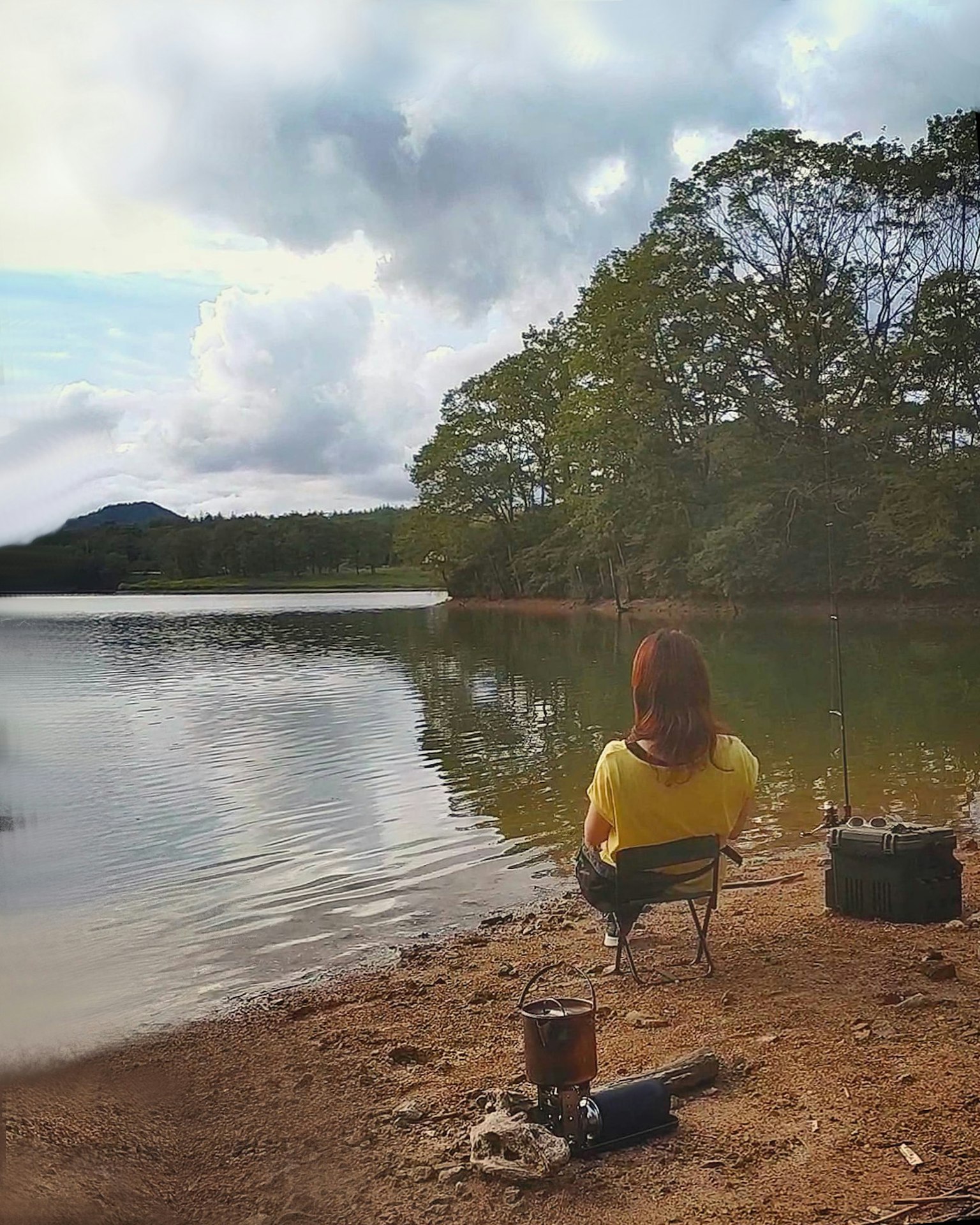 Frau, die am See angelt, trägt ein gelbes Shirt ruhiges Wasser und grüne Bäume im Hintergrund