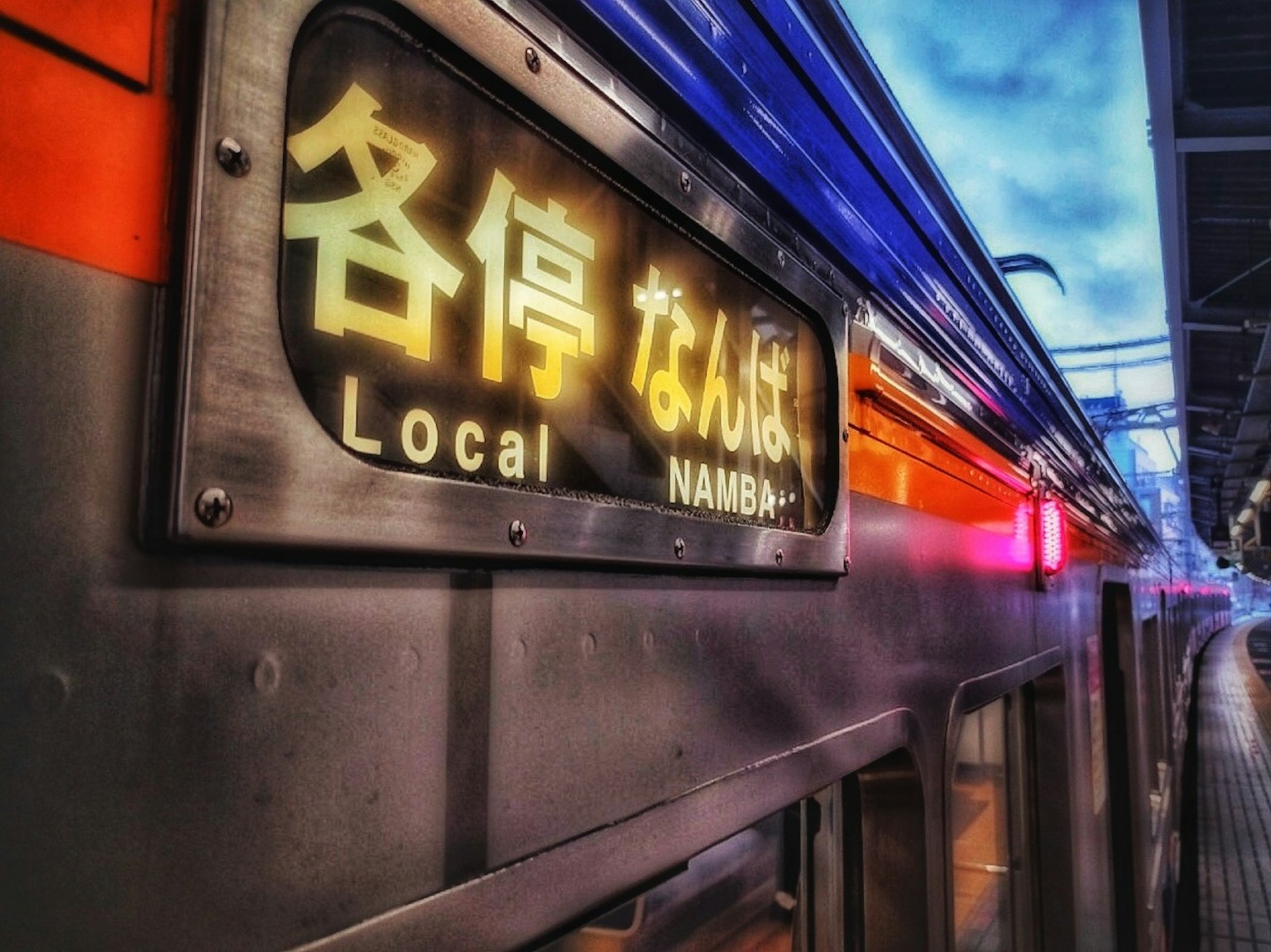 Papan kereta yang menampilkan Namba Local dengan suasana stasiun malam yang hidup