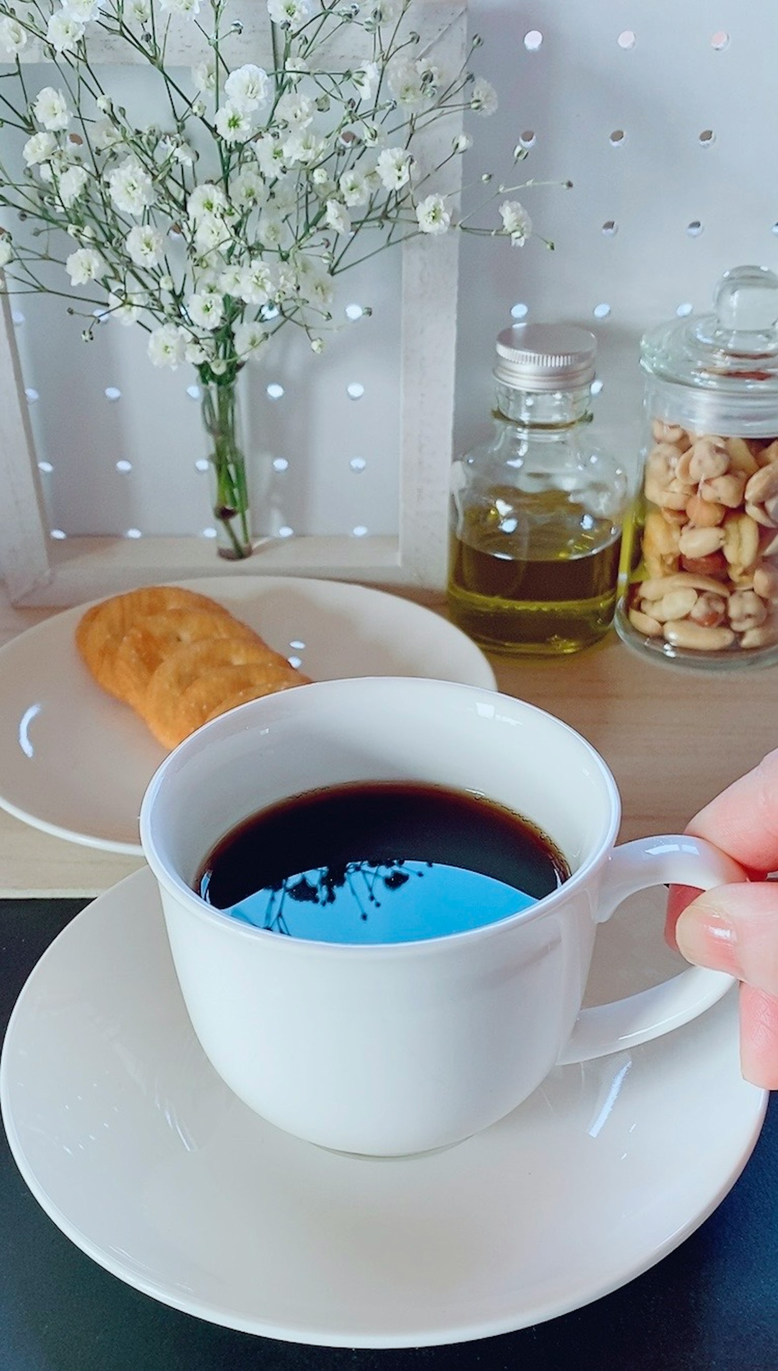 Một bàn tay cầm cốc cà phê trắng với cà phê đen bên cạnh đĩa bánh ngọt và hoa trang trí