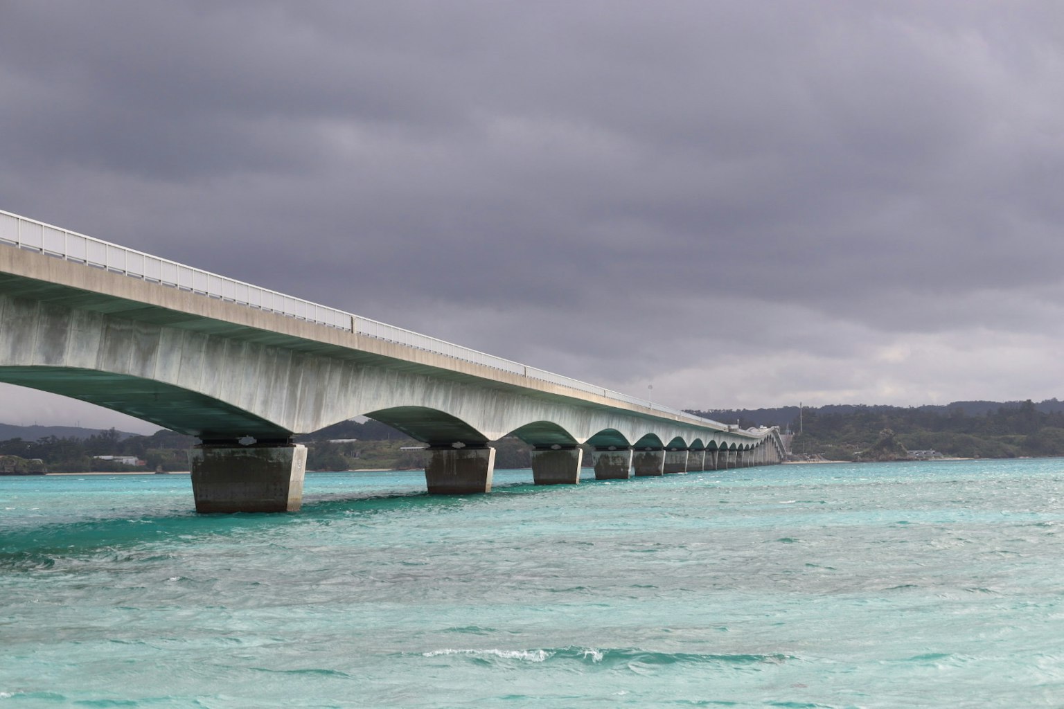 สะพานคอนกรีตเหนือทะเลสีฟ้าใสใต้ท้องฟ้าสีเทา