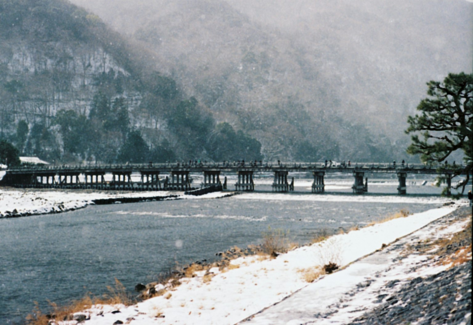 Schneebedeckte Flusslandschaft mit einer Brücke und Bergen im Hintergrund