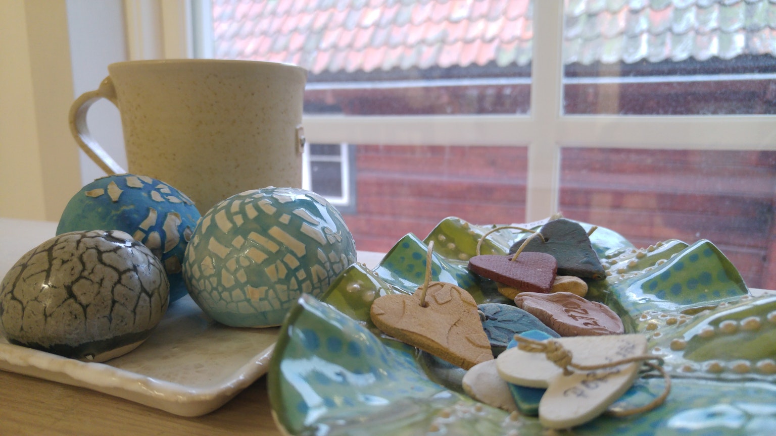 A ceramic cup and colorful stone objects on a table with a view of a red-roofed house through the window