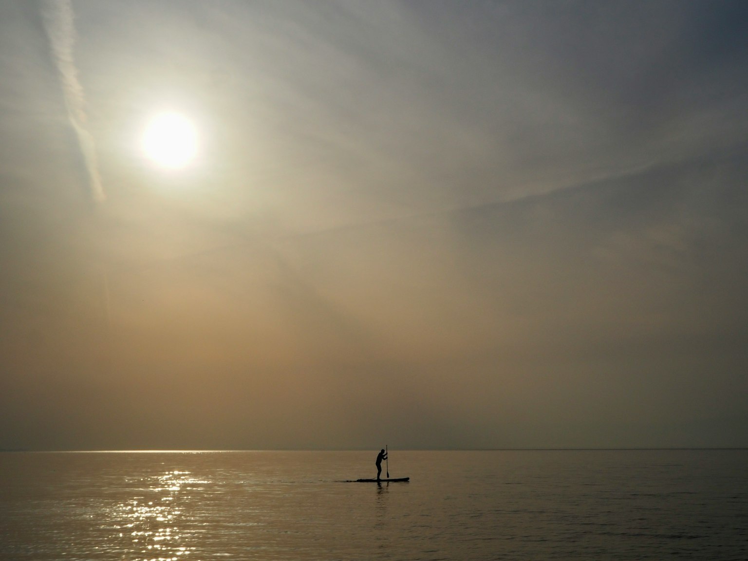 一个人在平静的海面上划着立式桨板,背景是明亮的太阳