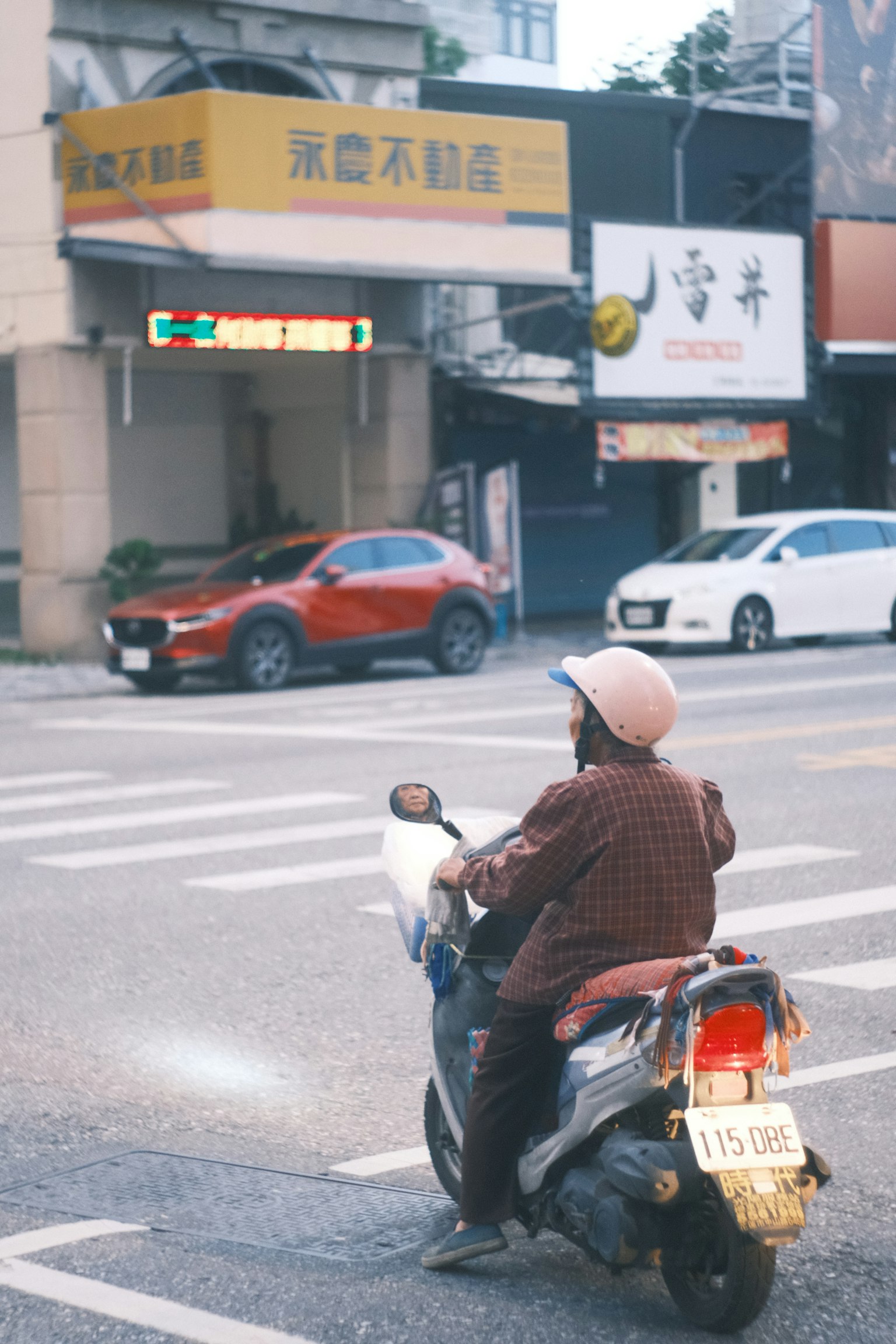 一個騎著滑板車的人正在過馬路，旁邊有商店和汽車