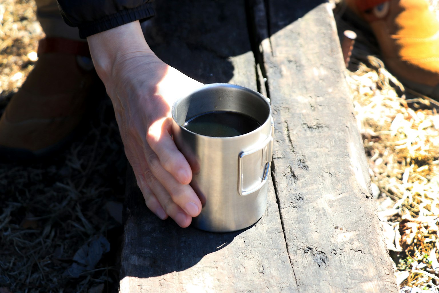 Una mano sosteniendo una taza de metal apoyada en una viga de madera con un pie cerca