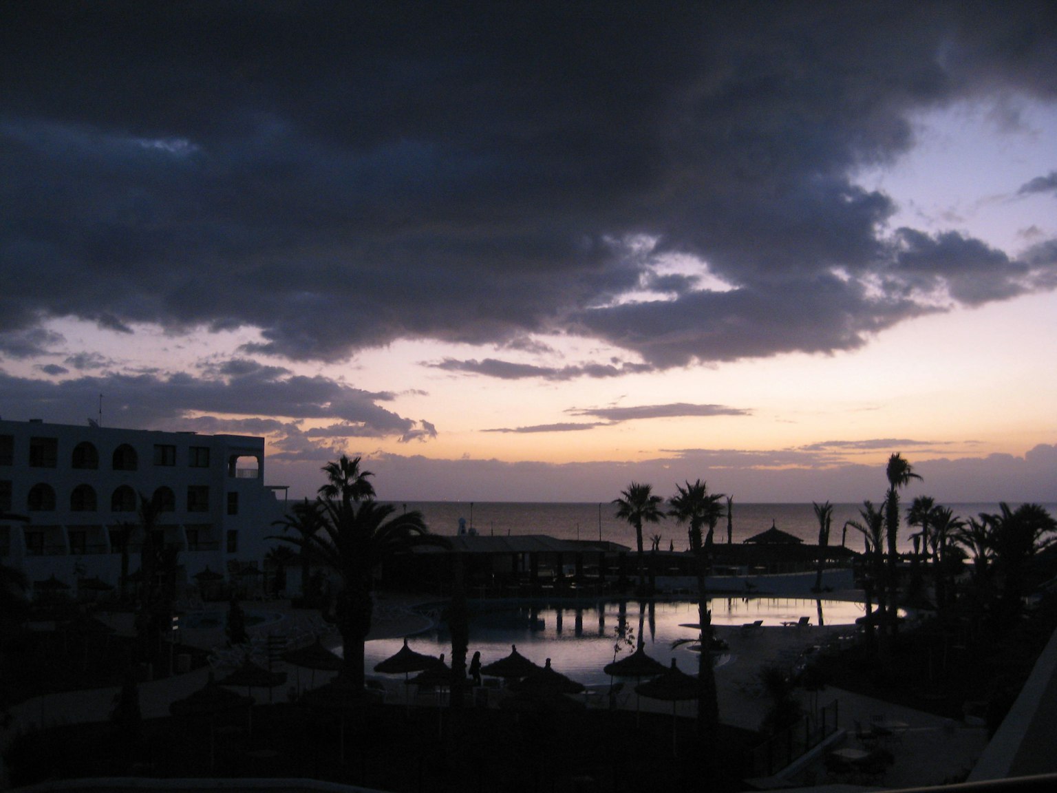 Vista al tramonto di un hotel resort e piscina con palme e nuvole scure sopra l'oceano