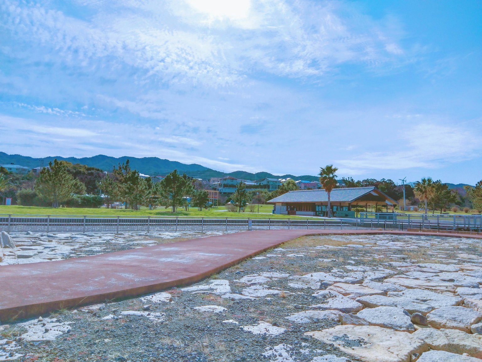 Pemandangan taman di bawah langit biru dengan gunung di latar belakang jalan yang dipaving dan pohon hijau