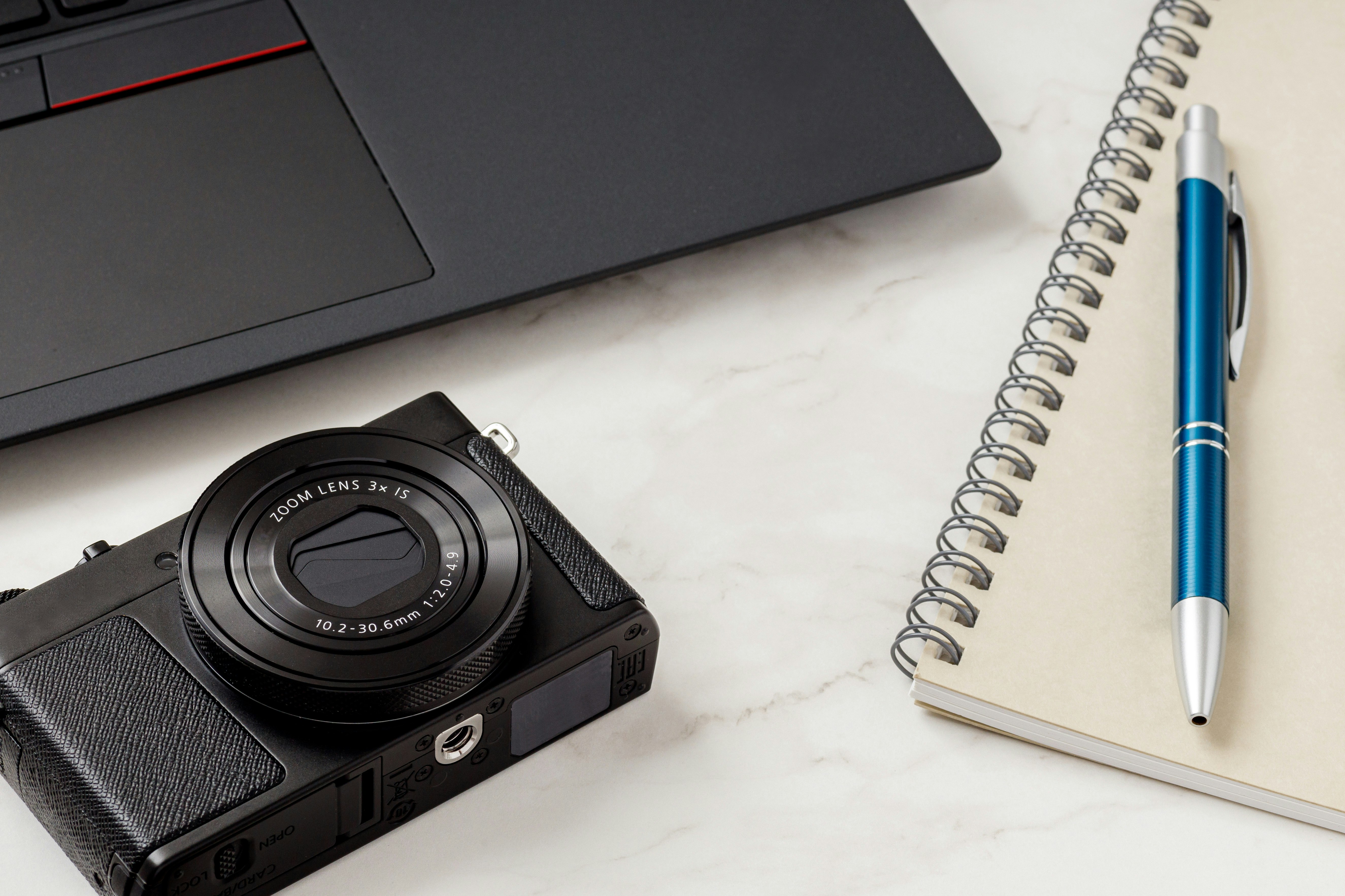 desk setup with a compact camera, notebook and pen beside a laptop