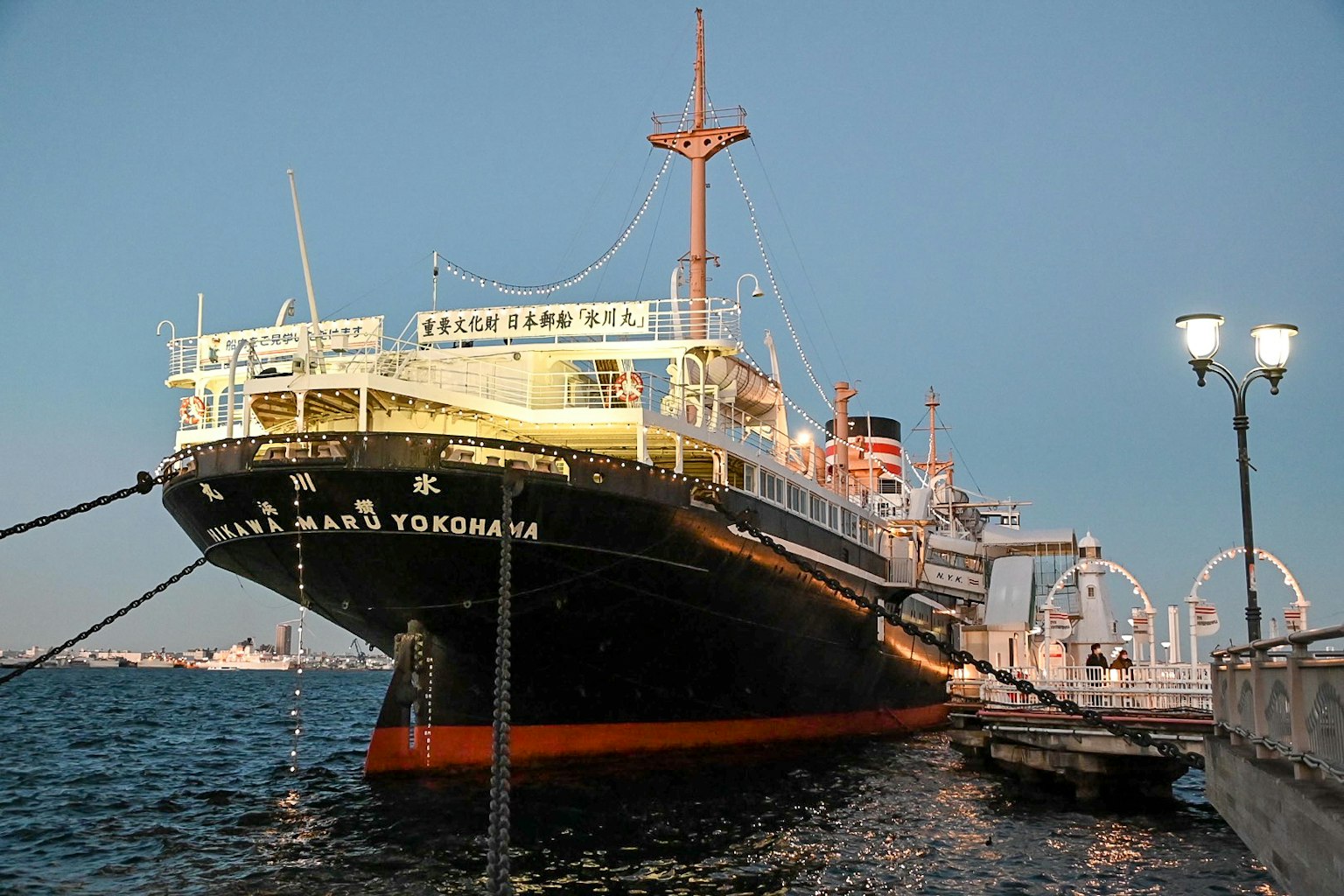 Image d'un navire historique amarré dans des eaux bleues Le navire s'appelle 'Nippon Maru' avec une coque noire et blanche illuminée