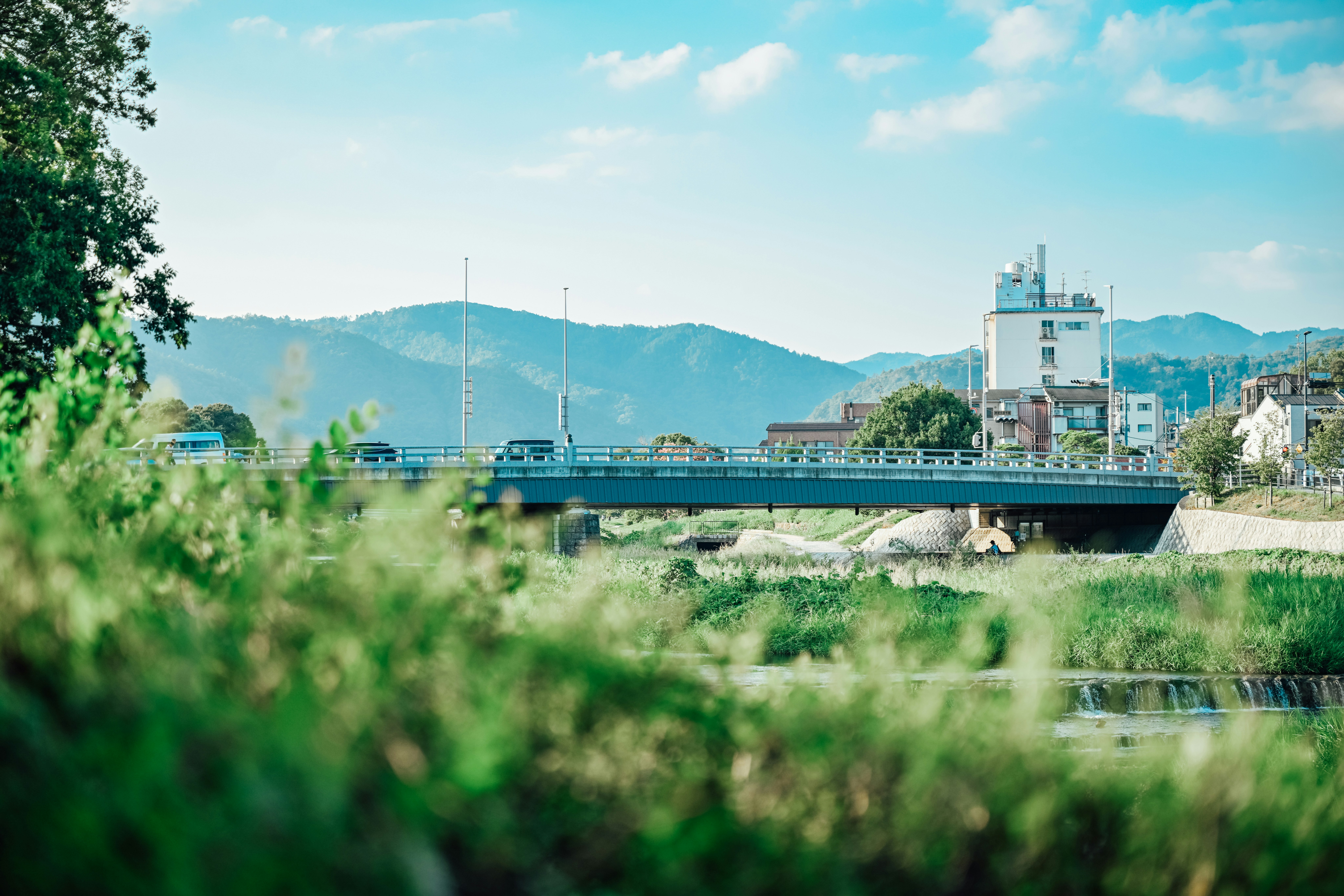 川沿いの緑の草むらと橋、遠くに工場の煙突が見える風景