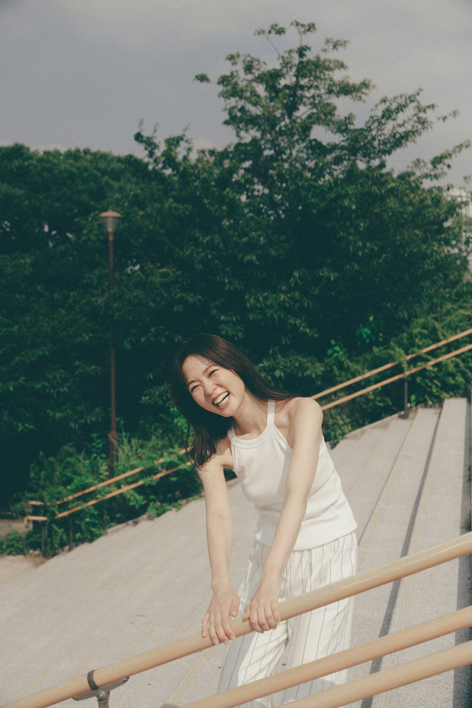 A woman in a white outfit leans on a railing smiling with greenery in the background