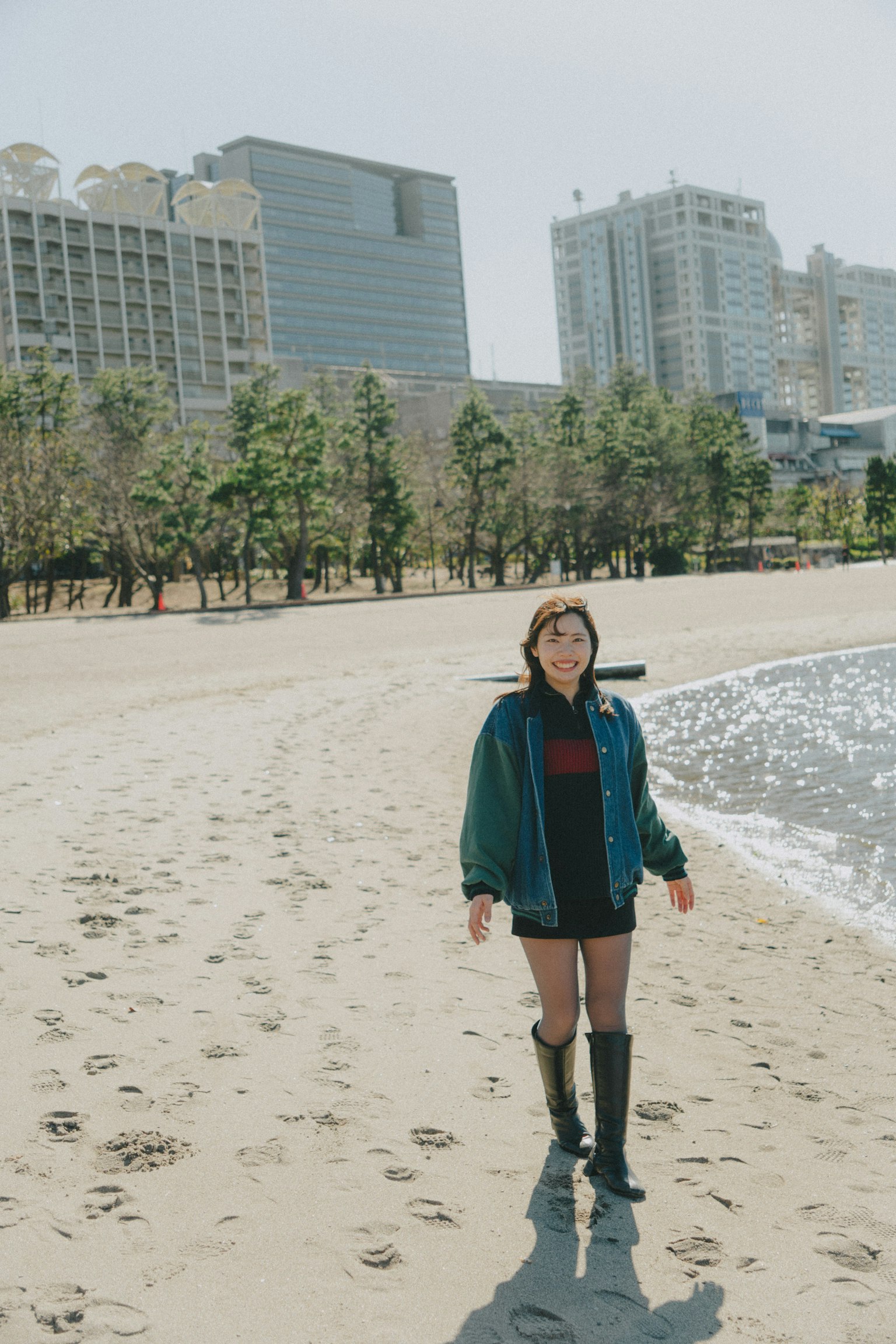 Mujer de pie en la playa con chaqueta azul y pantalones cortos negros rodeada de edificios altos y árboles