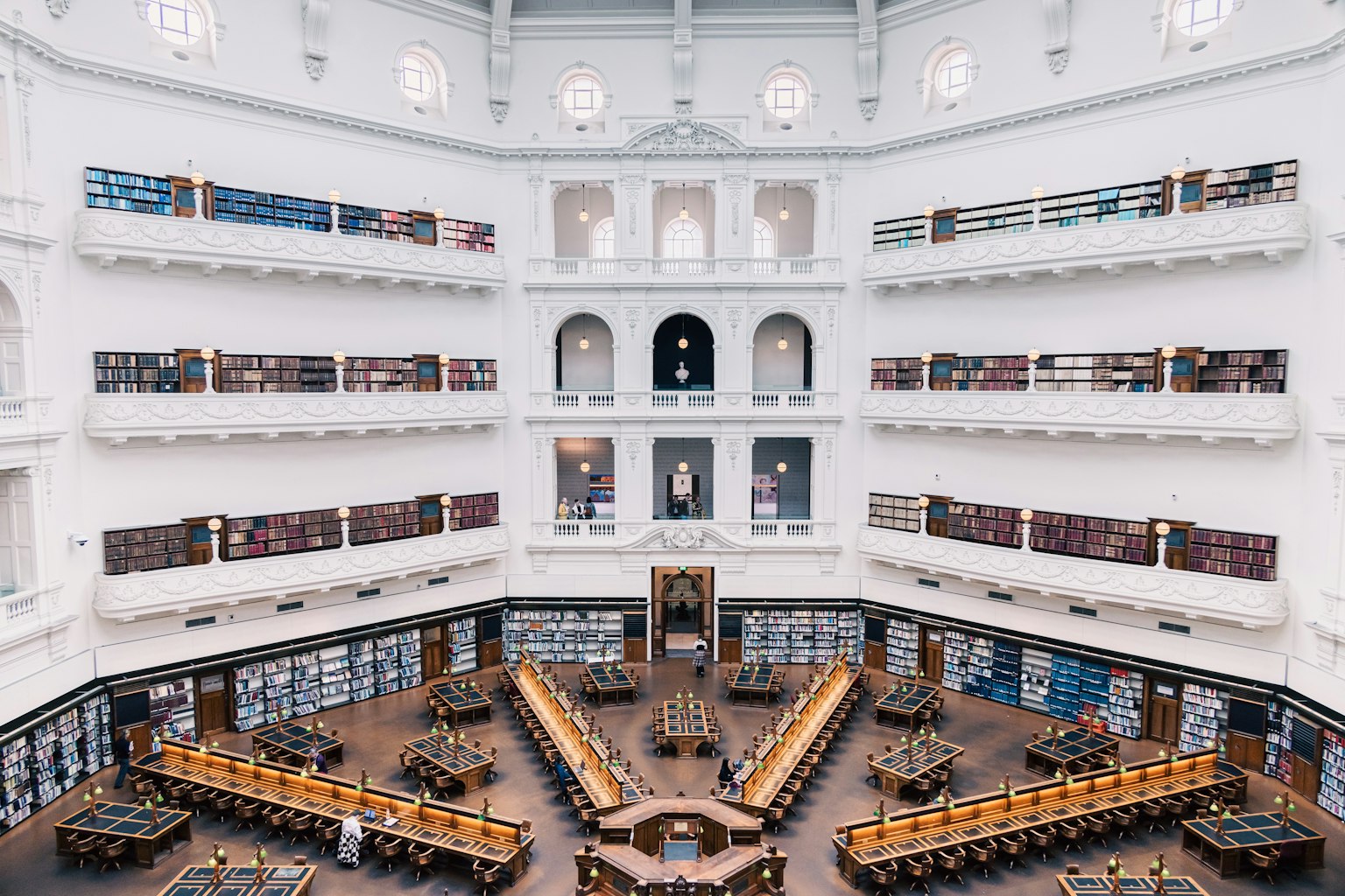 ภายในห้องสมุดที่สวยงาม มีผนังสีขาวและโต๊ะไม้ล้อมรอบด้วยชั้นวางหนังสือ