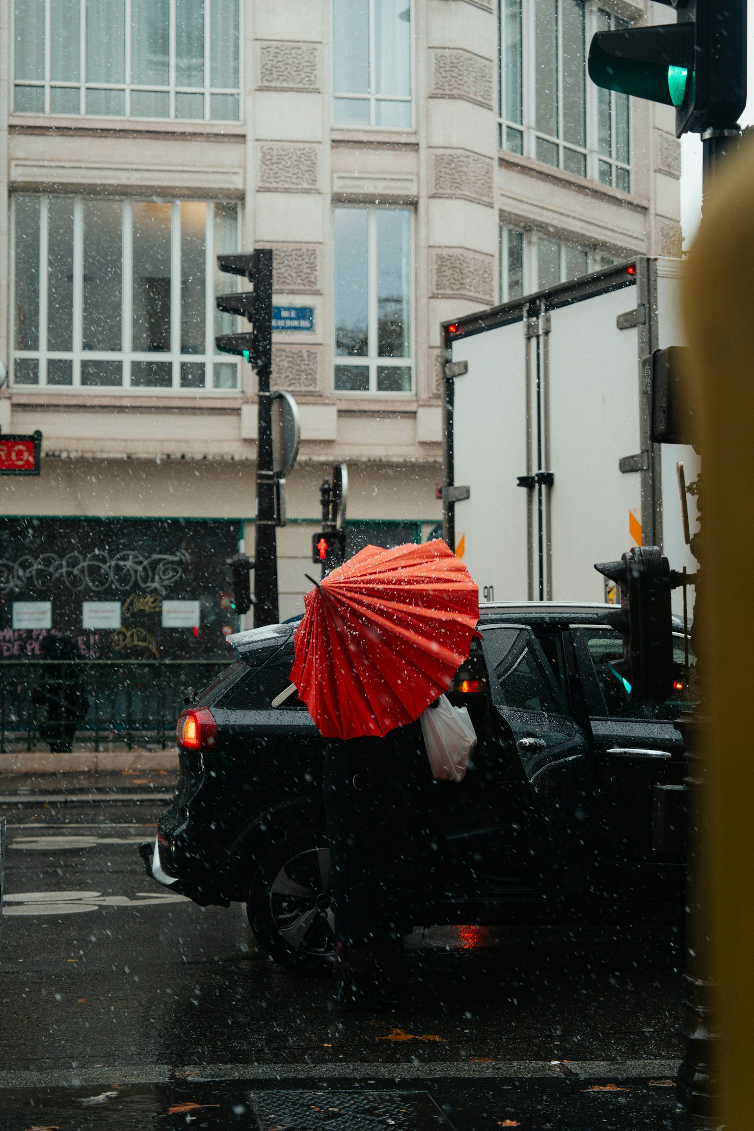 Una persona con un ombrello rosso si trova accanto a un'auto nera sotto la pioggia