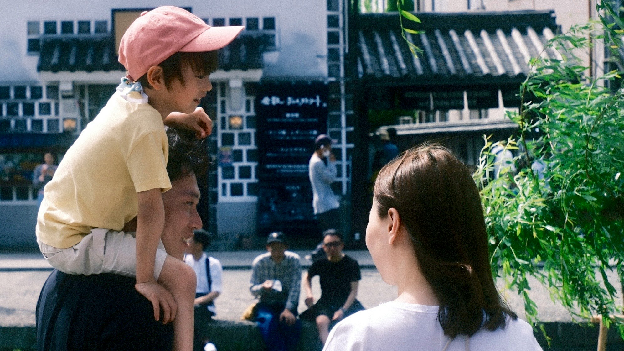 公園の緑のそばで帽子をかぶった子どもと大人が見つめ合う日常のひととき