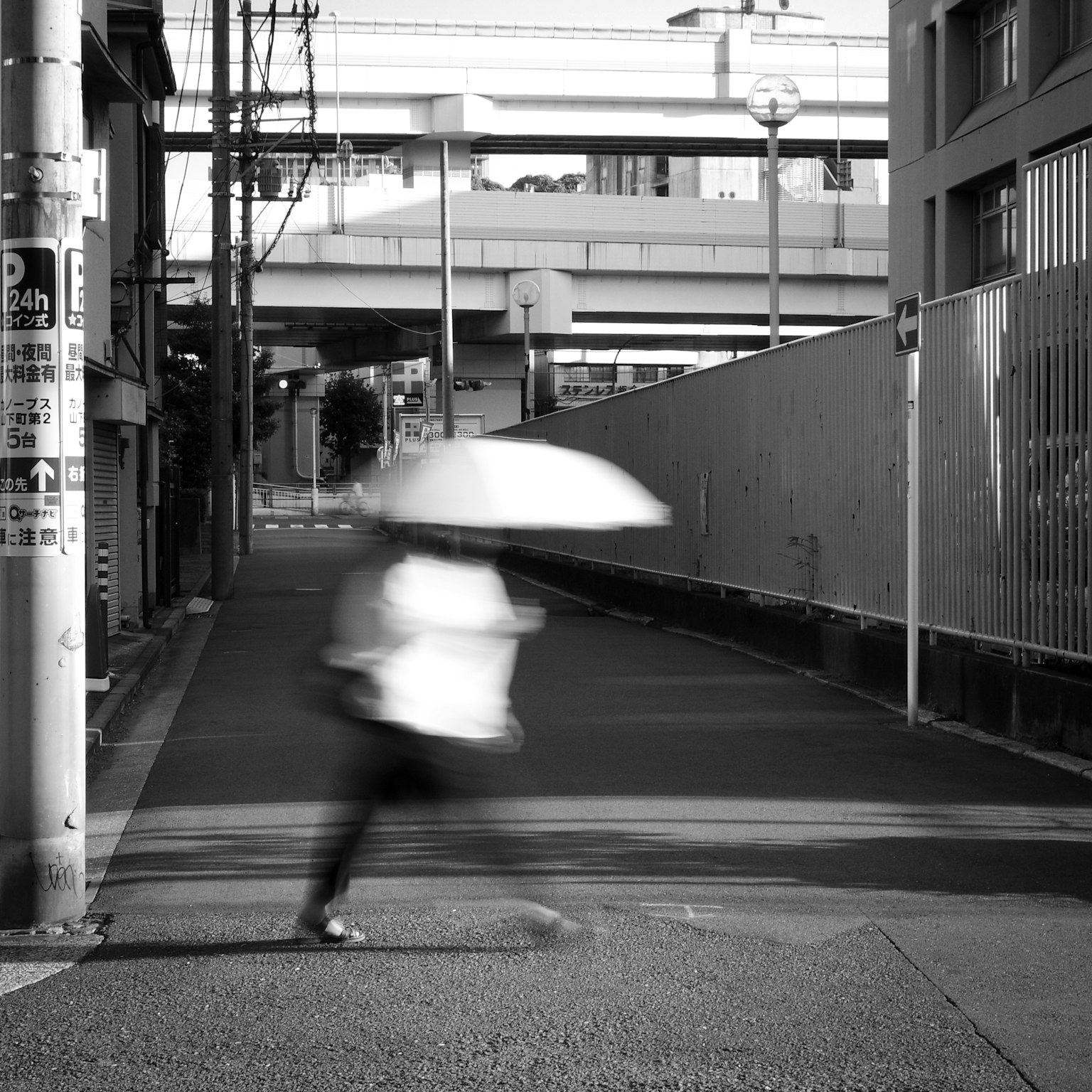 Une silhouette floue marchant avec un parapluie dans une rue en noir et blanc avec une route surélevée