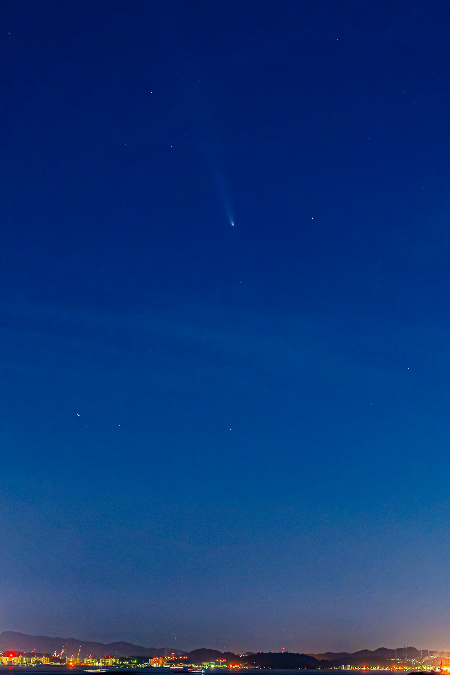 Un cometa visible en el cielo nocturno con estrellas y un horizonte urbano abajo