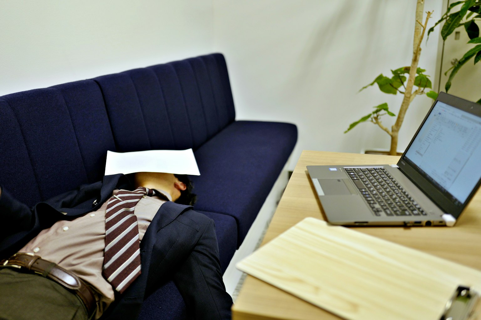 Doanh nhân ngủ trên ghế sofa màu xanh với laptop và tài liệu gần đó