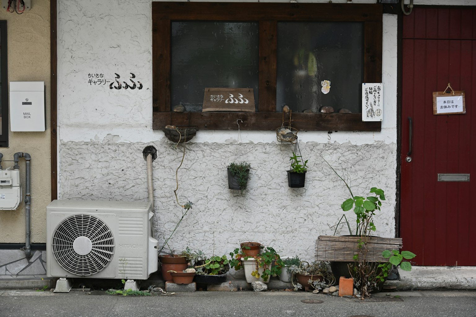 Exterior of a shop with white walls and a red door featuring plants on the window and an air conditioning unit
