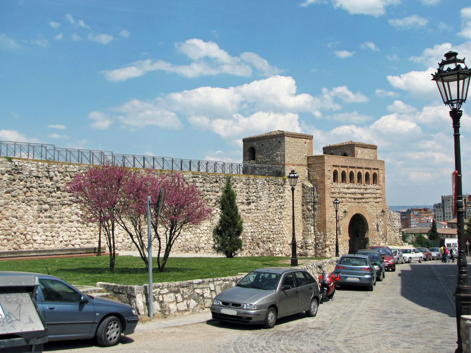 Vista panoramica di un antico muro di pietra e di un cancello con ciliegi in fiore e auto parcheggiate