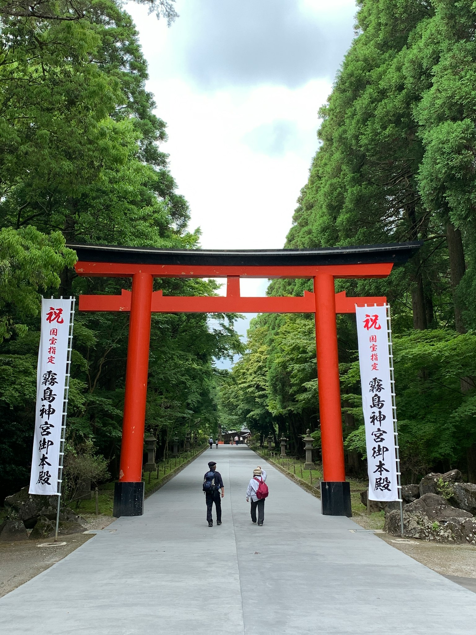 Due persone che camminano sotto un torii rosso circondato da una vegetazione lussureggiante