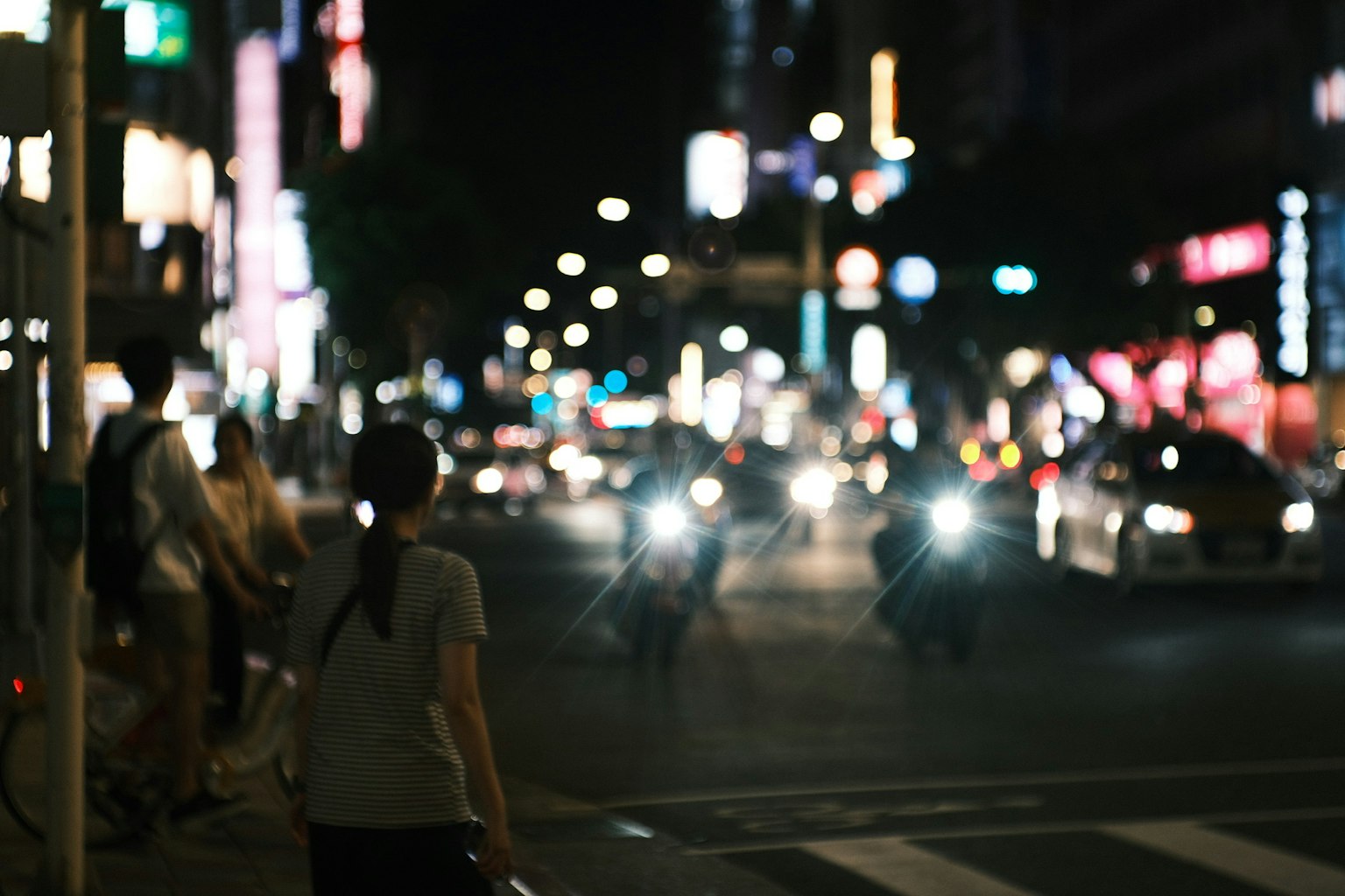 Pejalan kaki menunggu di persimpangan kota di malam hari dengan lampu depan mobil bersinar dan papan iklan neon terlihat