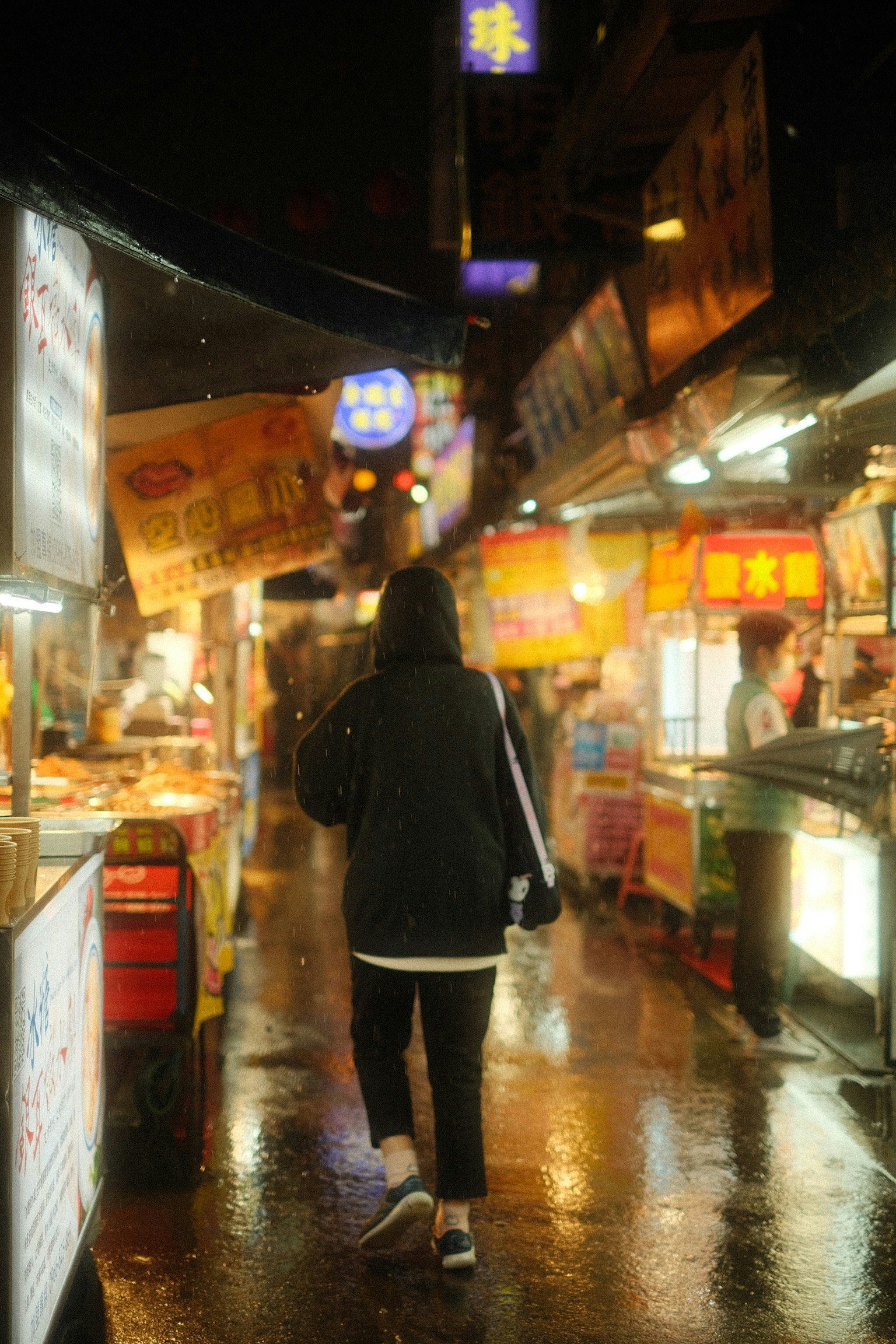 Personne marchant dans un marché nocturne avec des stands colorés et des reflets sur le pavé mouillé