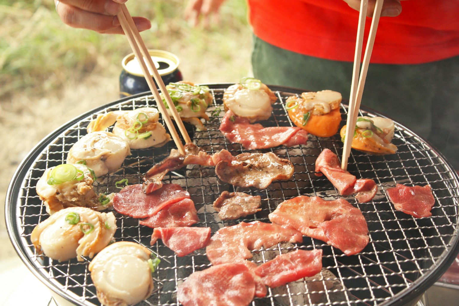 Person grilling various meats and seafood using chopsticks on a barbecue grill