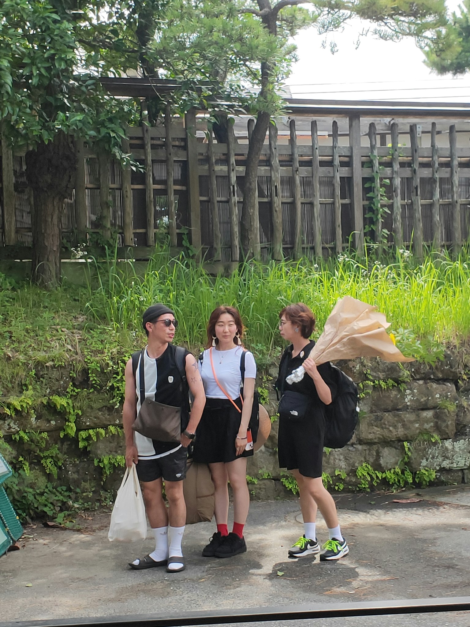 Three young people standing by the roadside with green grass and trees in the background