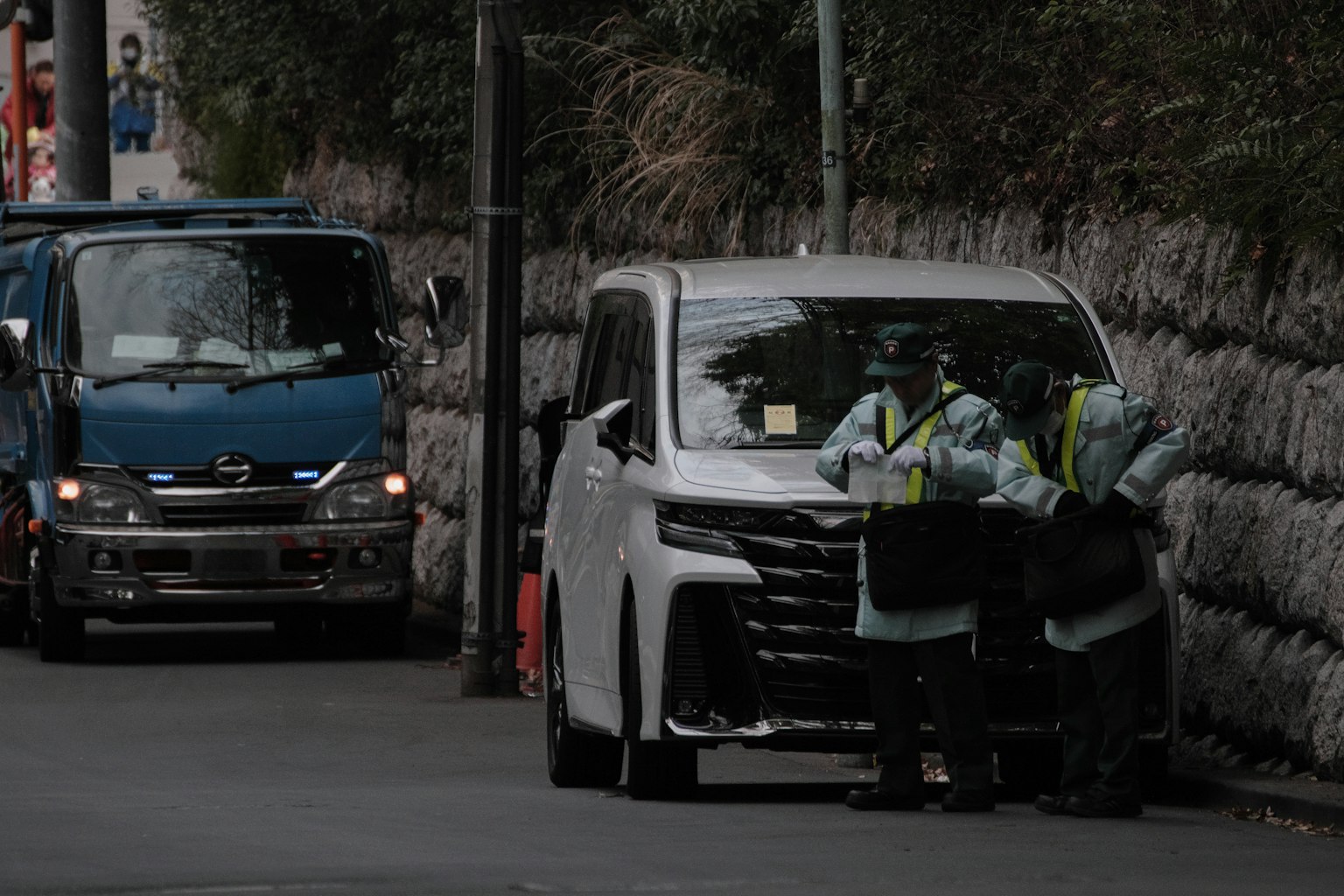 Two security personnel working near a white vehicle and a blue truck on the road