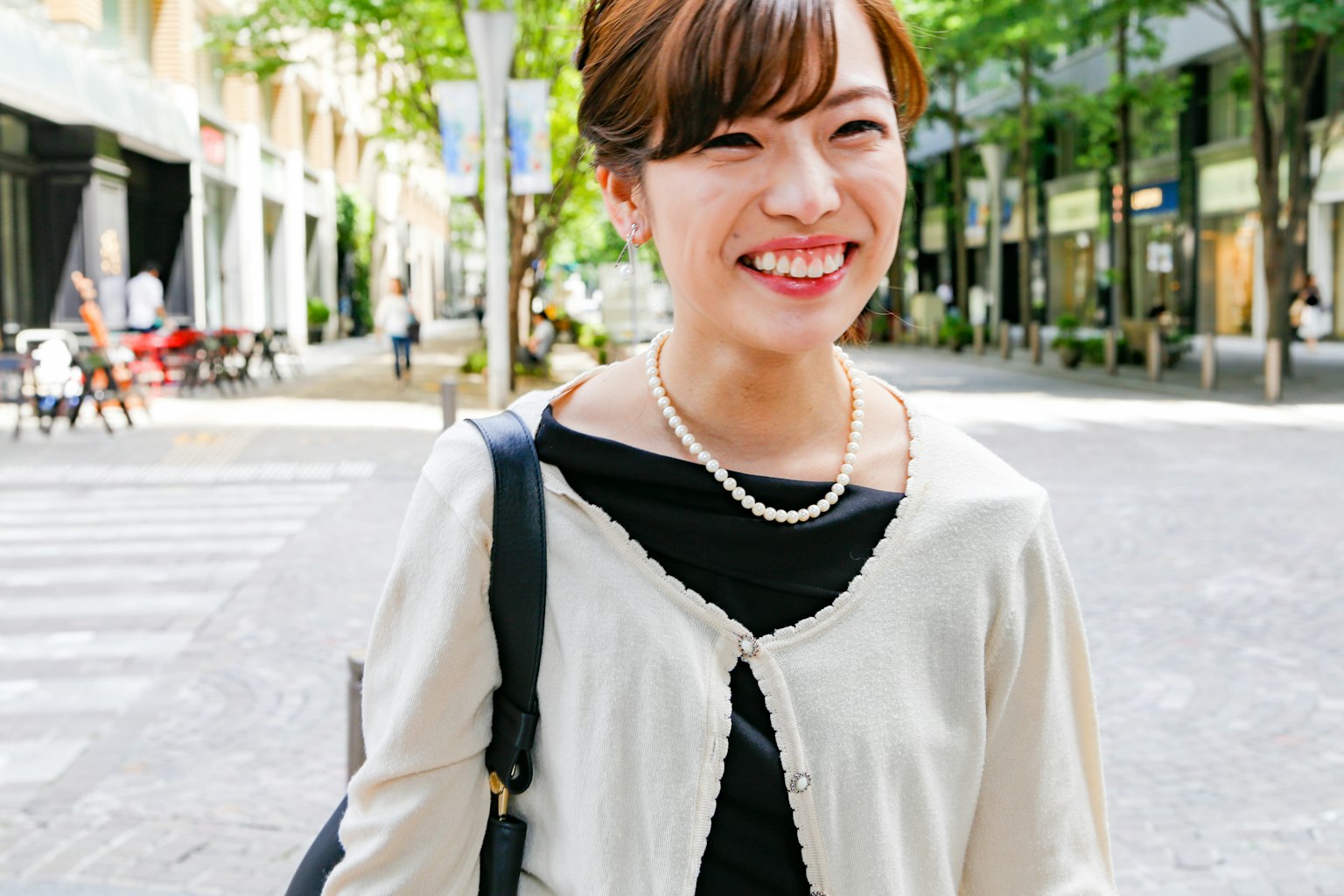 A smiling woman standing in the street with green trees and shops in the background